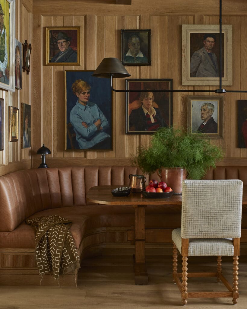 Cozy dining nook with a brown leather banquette, wooden paneled walls, and multiple framed portraits; a table with fruit and a copper pitcher nearby.