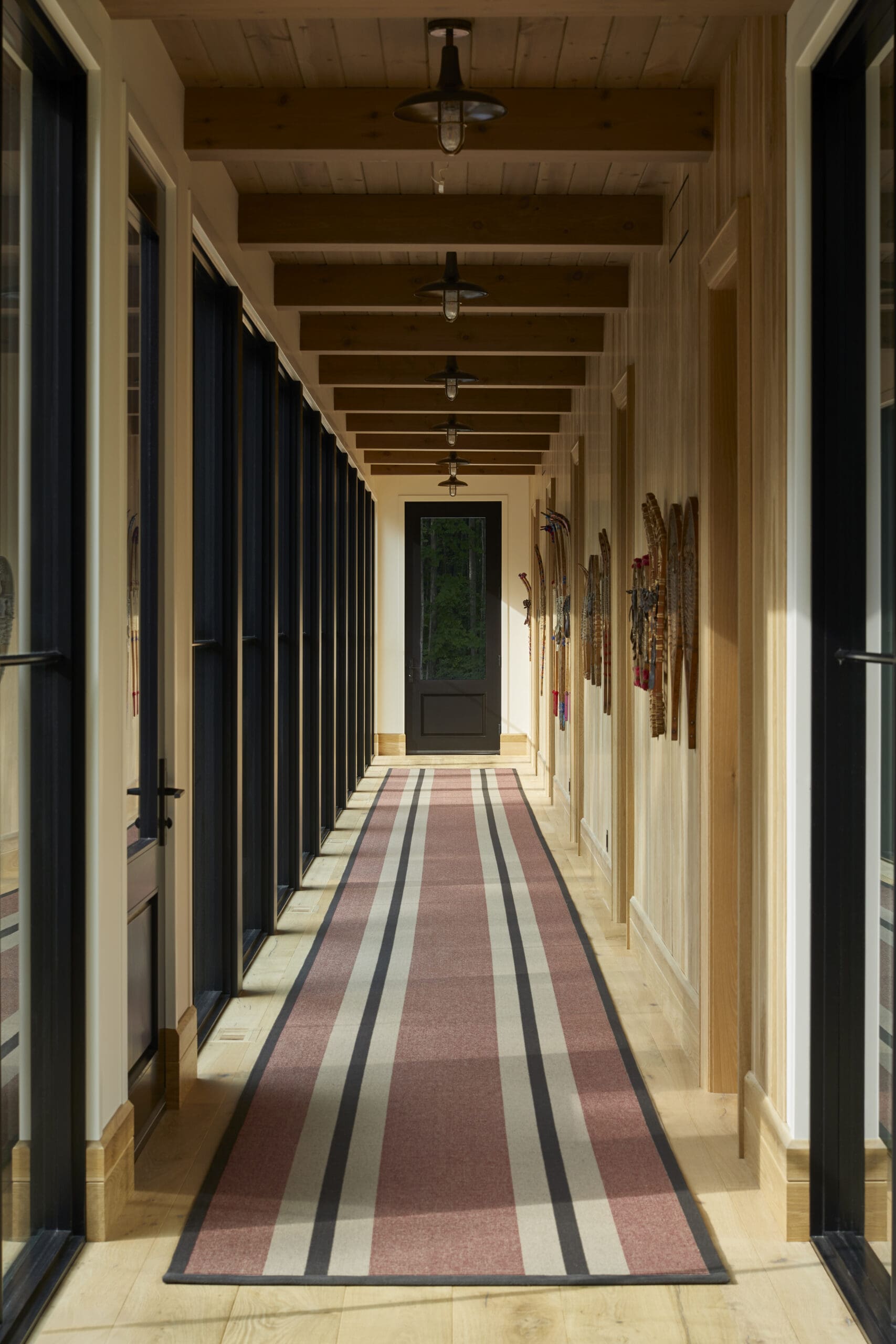 Long corridor with a red, gray, and beige striped rug, exposed wooden ceiling beams, and doors along the left side.
