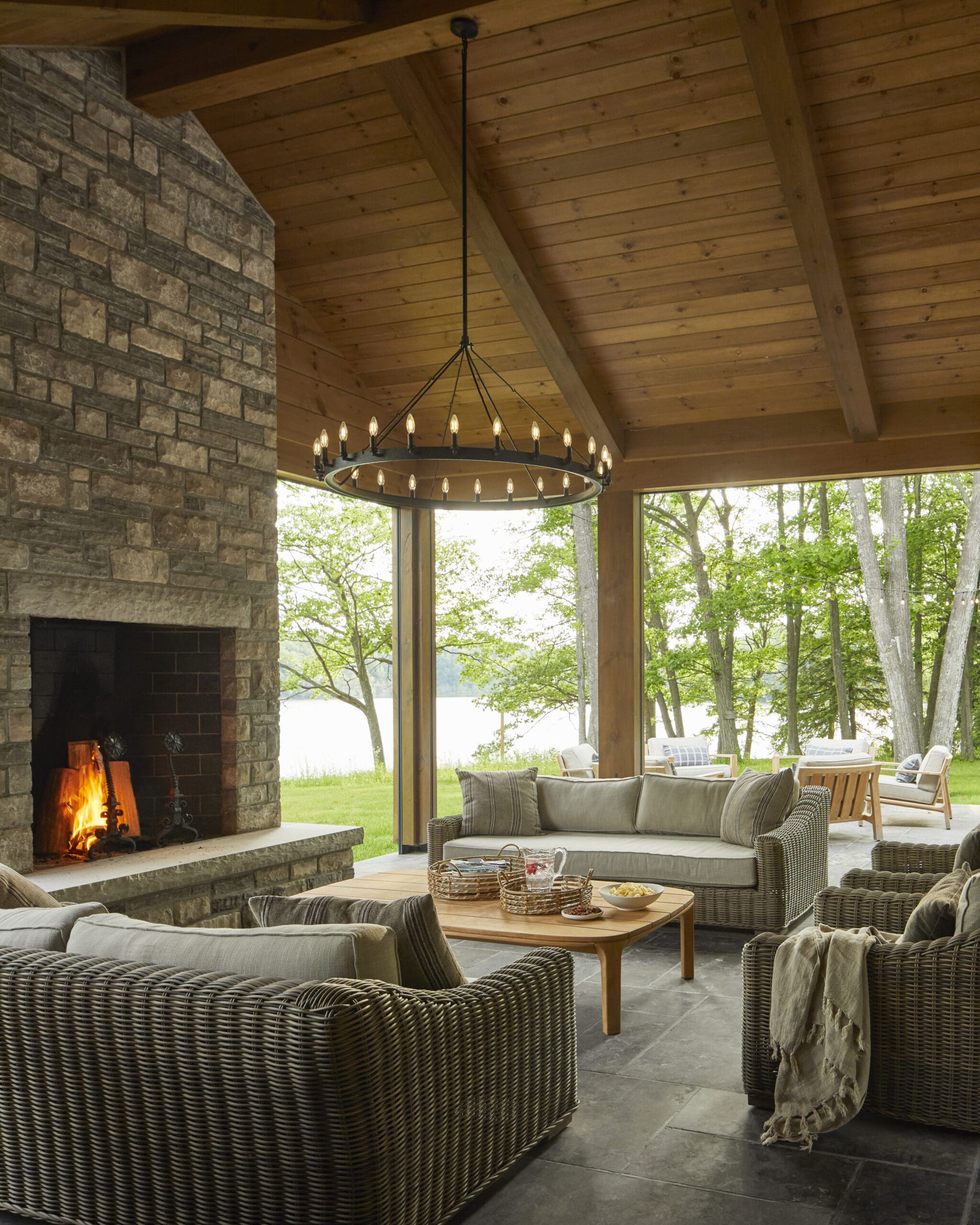 Cozy outdoor living space under a wood-paneled ceiling with a stone fireplace and a circular chandelier, wicker furniture, and a view of trees outside