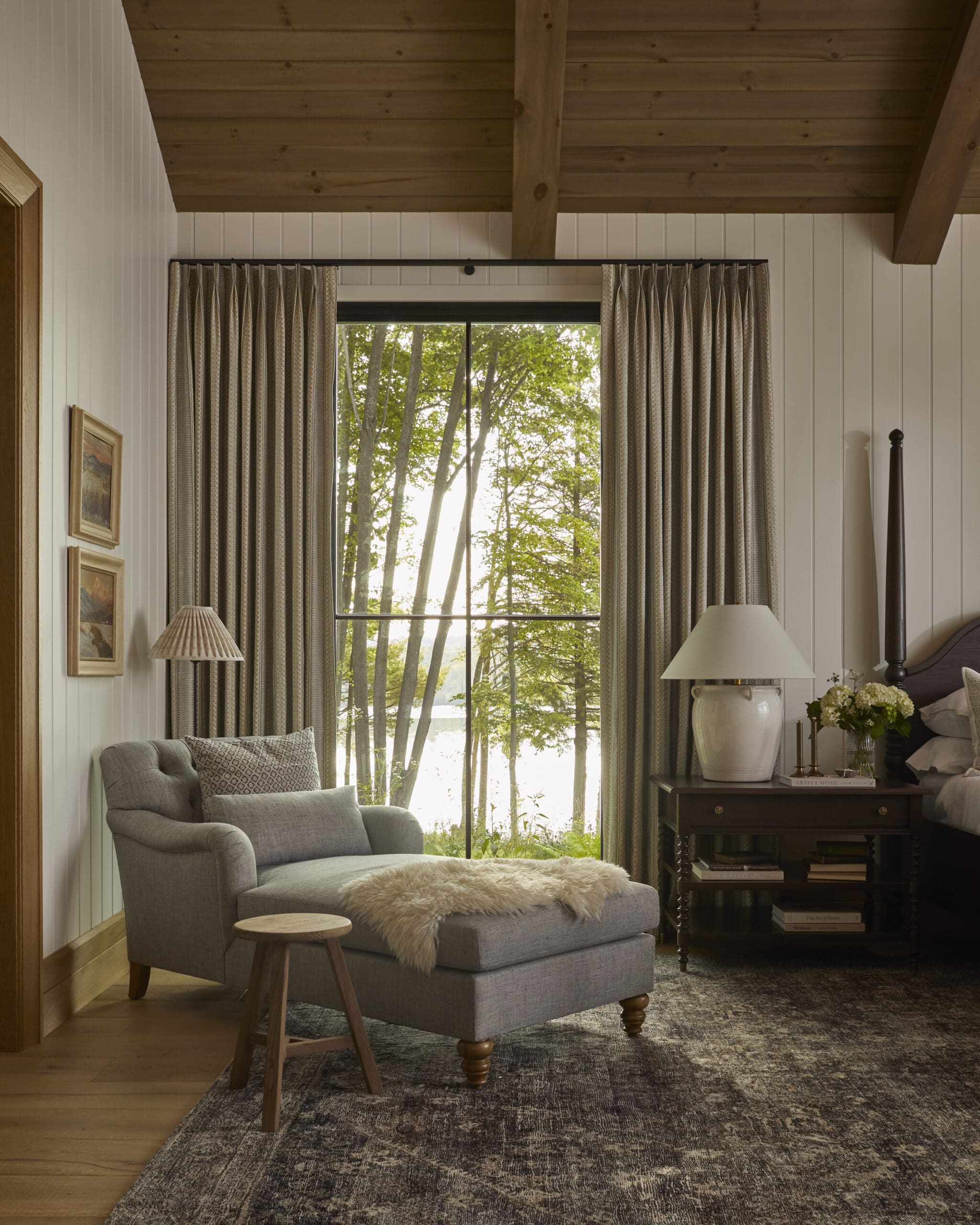 Cozy living room corner with a large window, beige patterned curtains, and a gray upholstered armchair beside a wooden side table and lamp.