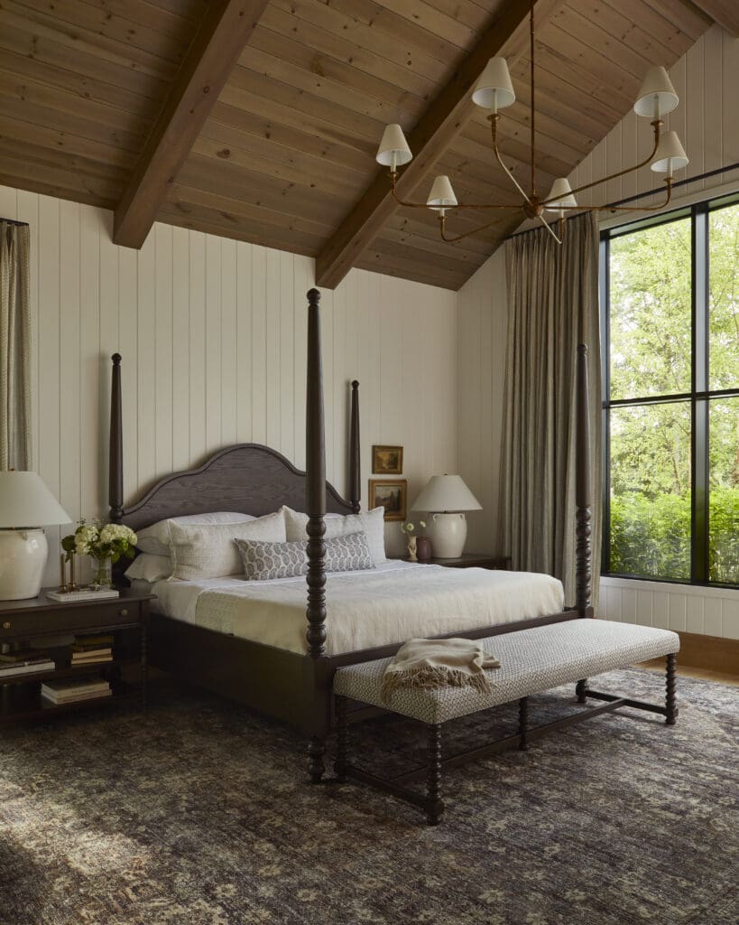 Rustic bedroom with a dark wooden four-poster bed, wood-paneled vaulted ceiling, and a large window with beige curtains.