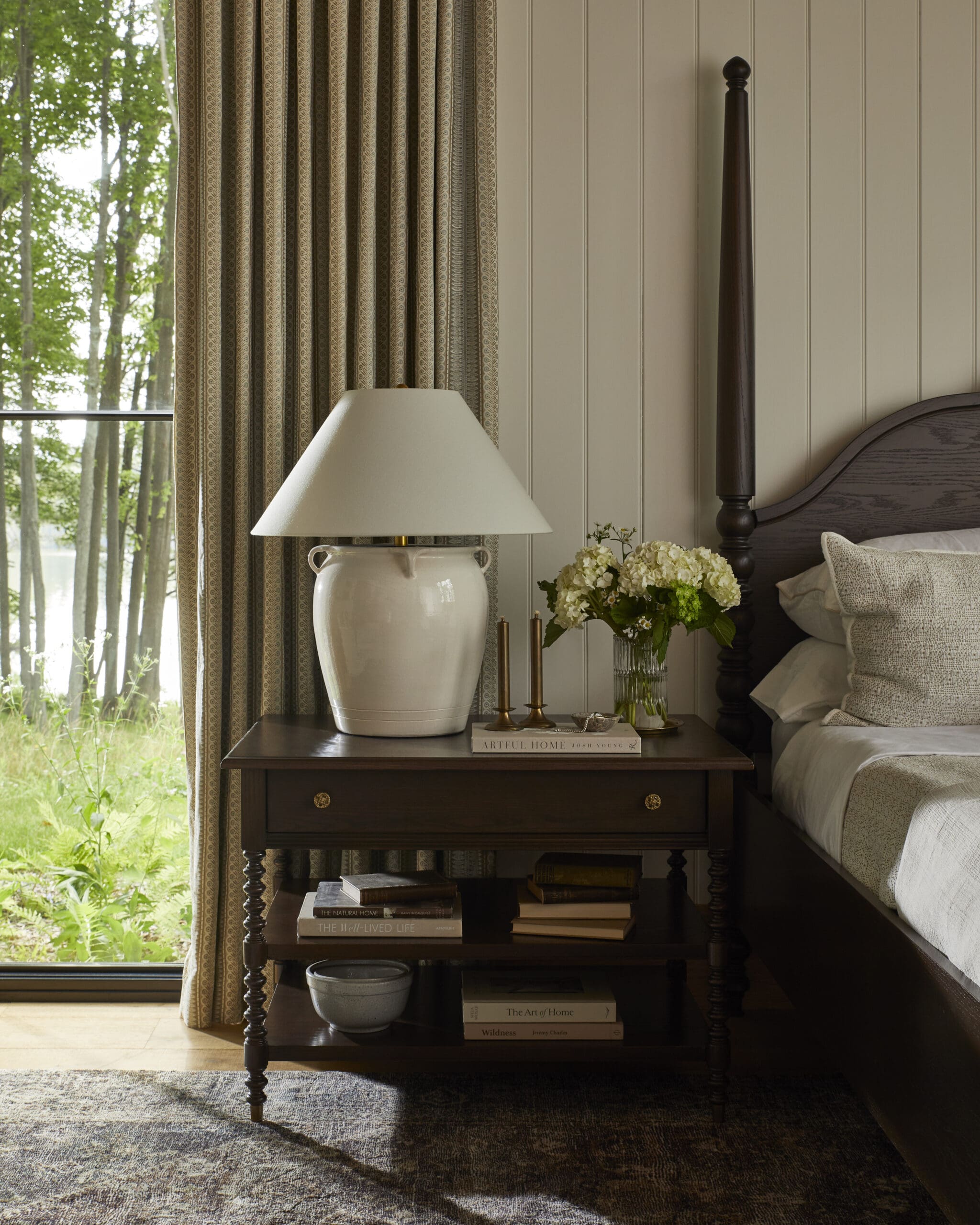 Cozy bedroom corner with a dark wooden nightstand, white ceramic lamp, a vase of white hydrangeas, and stacked books beside a beige-bedding bed.