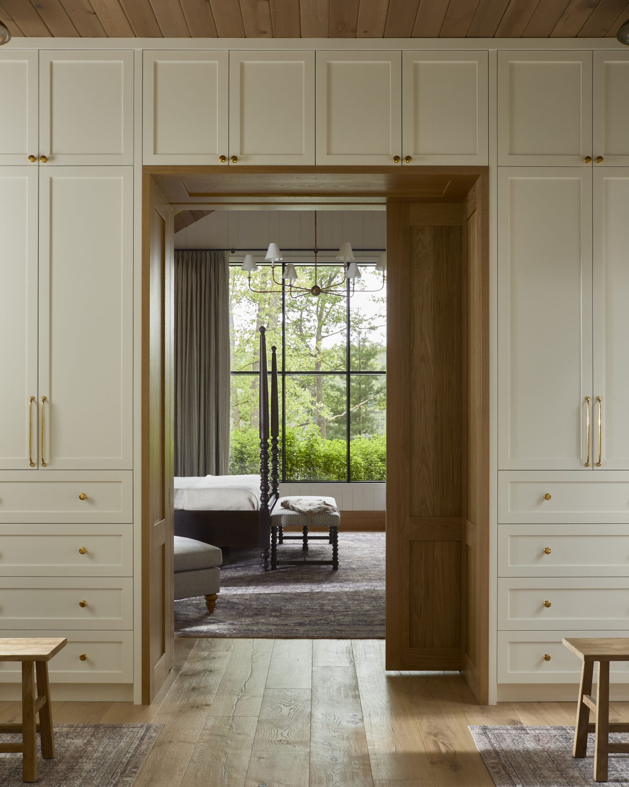 White built-in cabinets frame a wooden doorway leading into a bedroom with a four-poster bed and large window overlooking greenery.