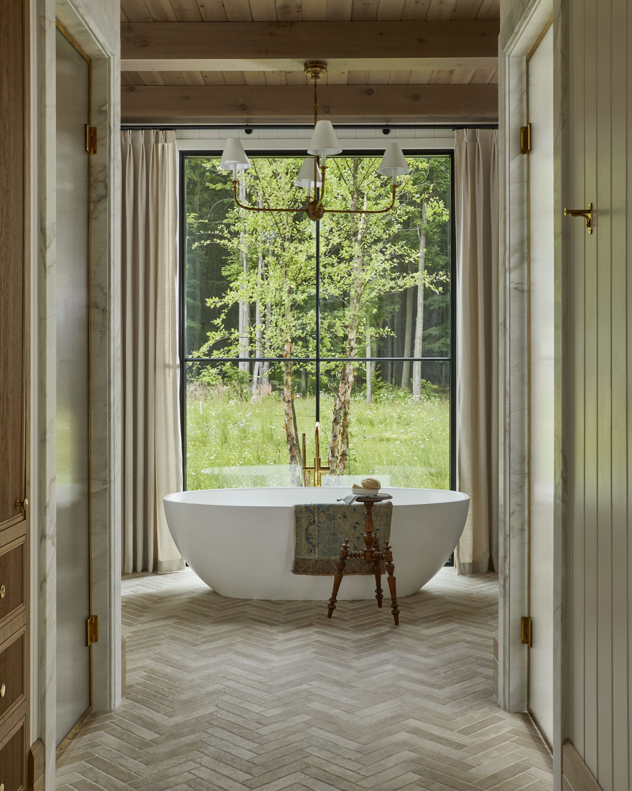 Freestanding white bathtub beneath a brass chandelier in a bright bathroom, with a large window framing green trees outside.