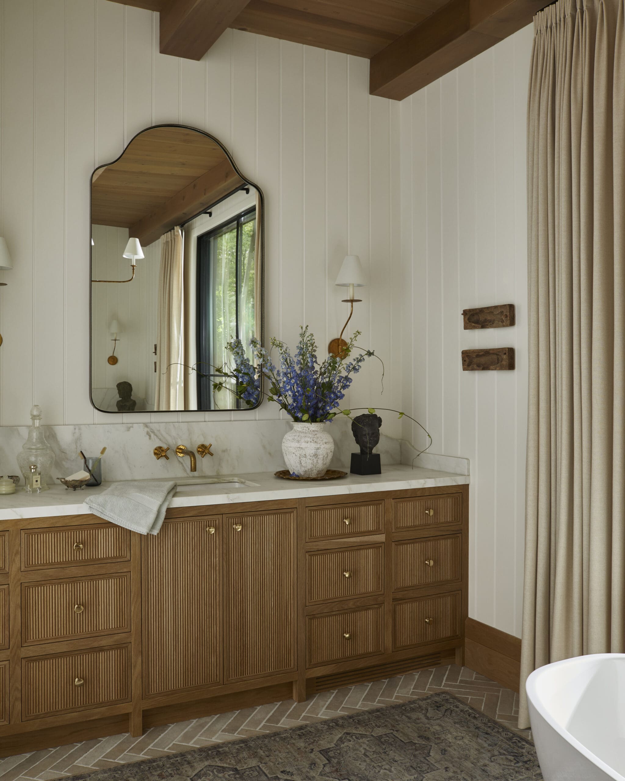 Wood-paneled bathroom with a large wooden vanity, marble countertop, and an arched mirror reflecting a window and greenery outside.