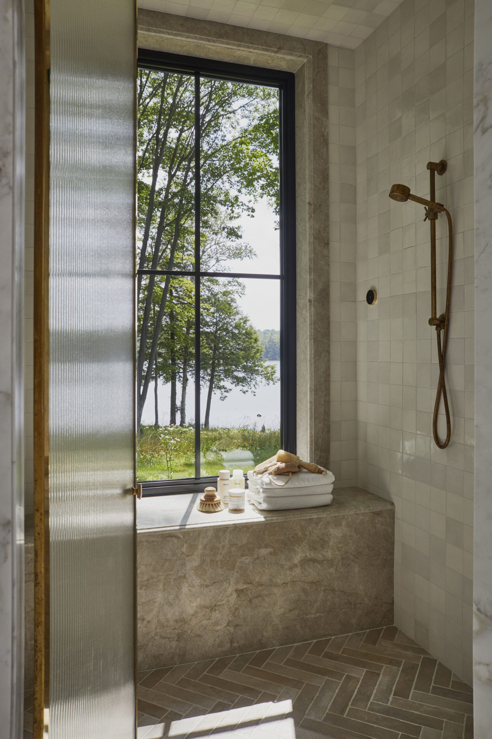 Shower area with brass handheld shower on tiled wall, window showing green trees outside and a bright outdoor view.