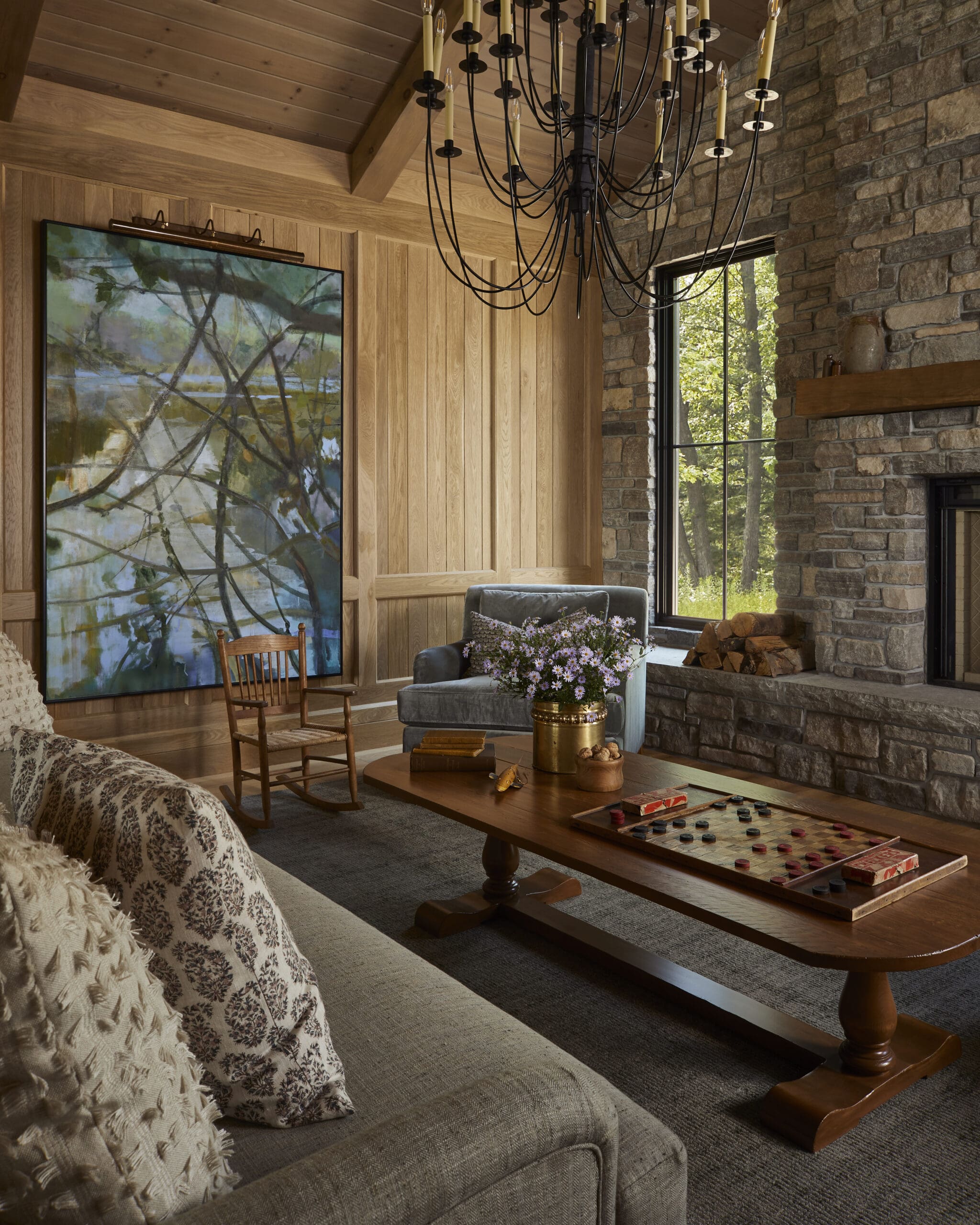 Cozy living room with a black metal chandelier, wood-paneled walls, and a stone fireplace; a large painting leans beside a wooden chair.