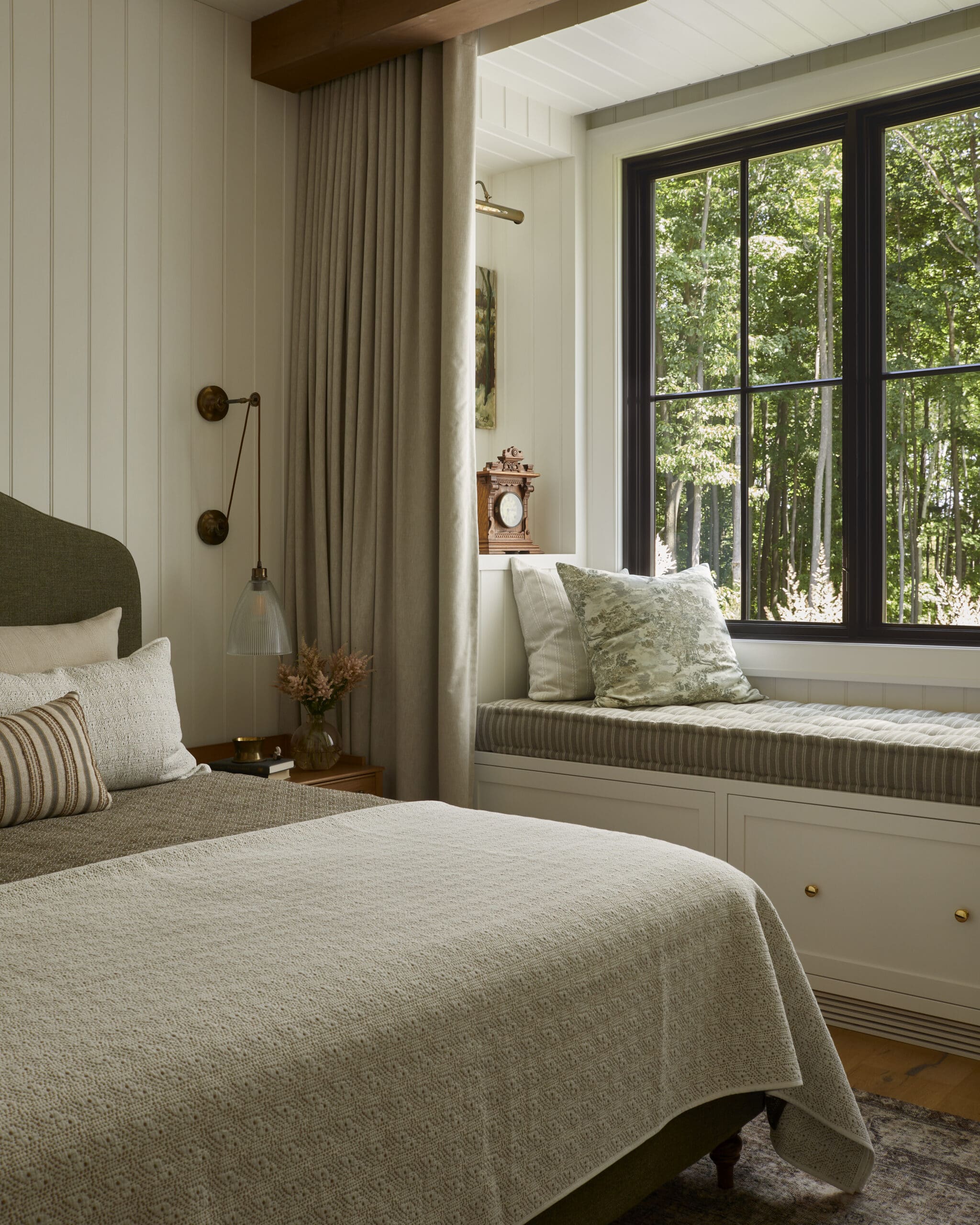 Cozy bedroom corner with a bed in cream bedding, decorative pillows, and a window seat by a large black-framed window showing green trees outside.