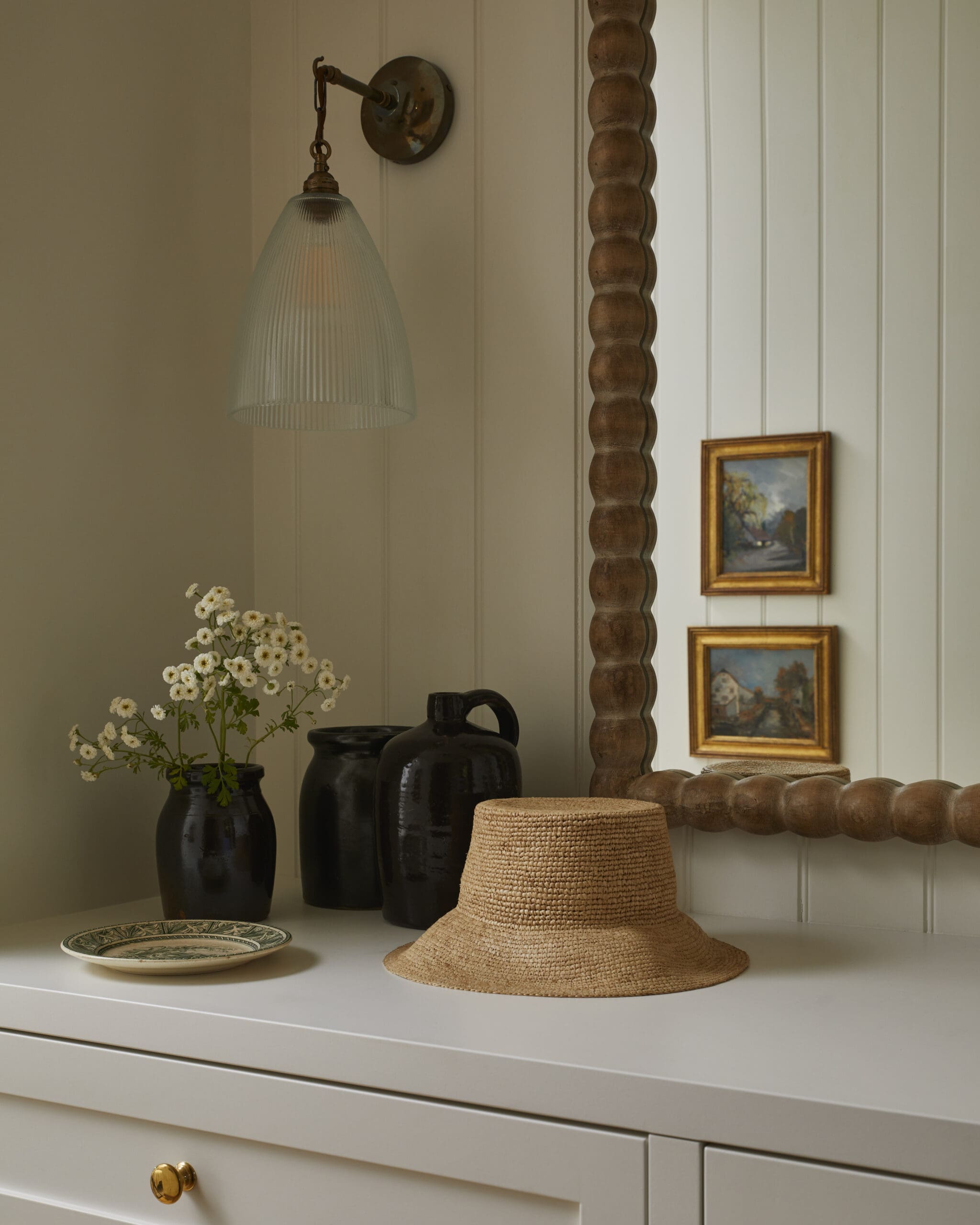 Cream cabinet scene featuring a frosted ribbed glass wall lamp, black ceramic vases, a woven straw hat, and small framed landscape paintings along a white paneled wall.