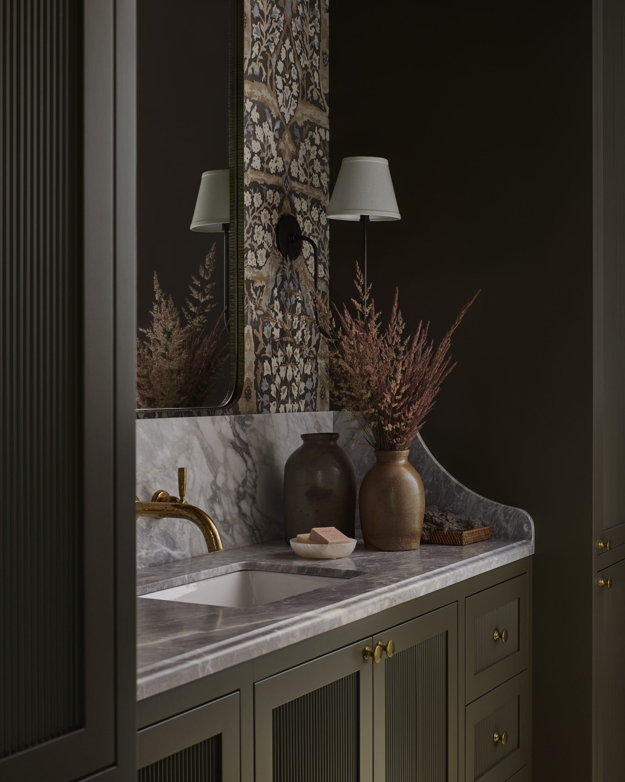 Dark bathroom vanity with marble countertop, patterned mirror panel, and two lamps casting warm light over decorative vases and soap dish in muted tones.
