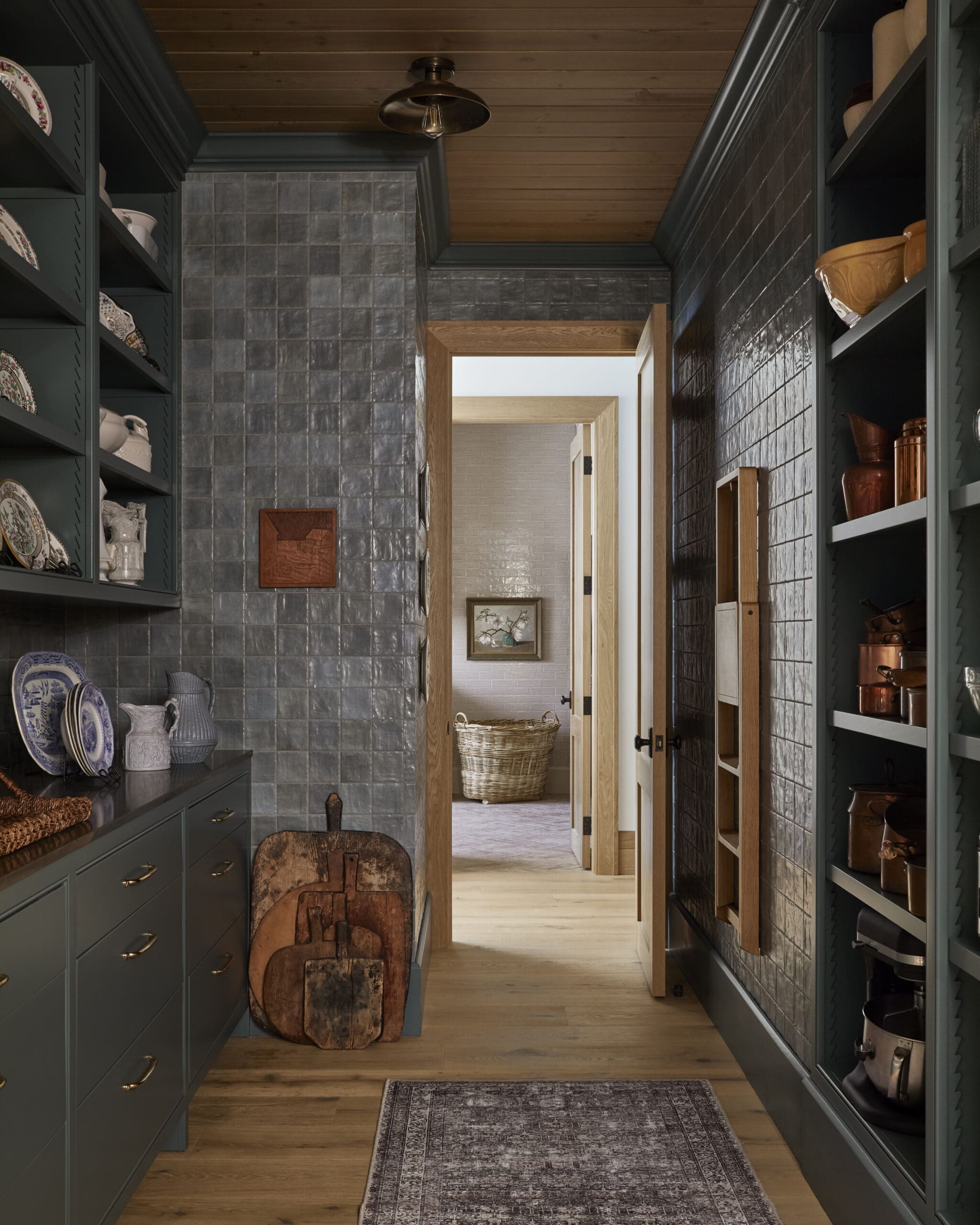 Narrow pantry with dark teal cabinetry and open shelves displaying plates and pottery, wooden ceiling, gray tiled walls, and a doorway to a sunlit room.