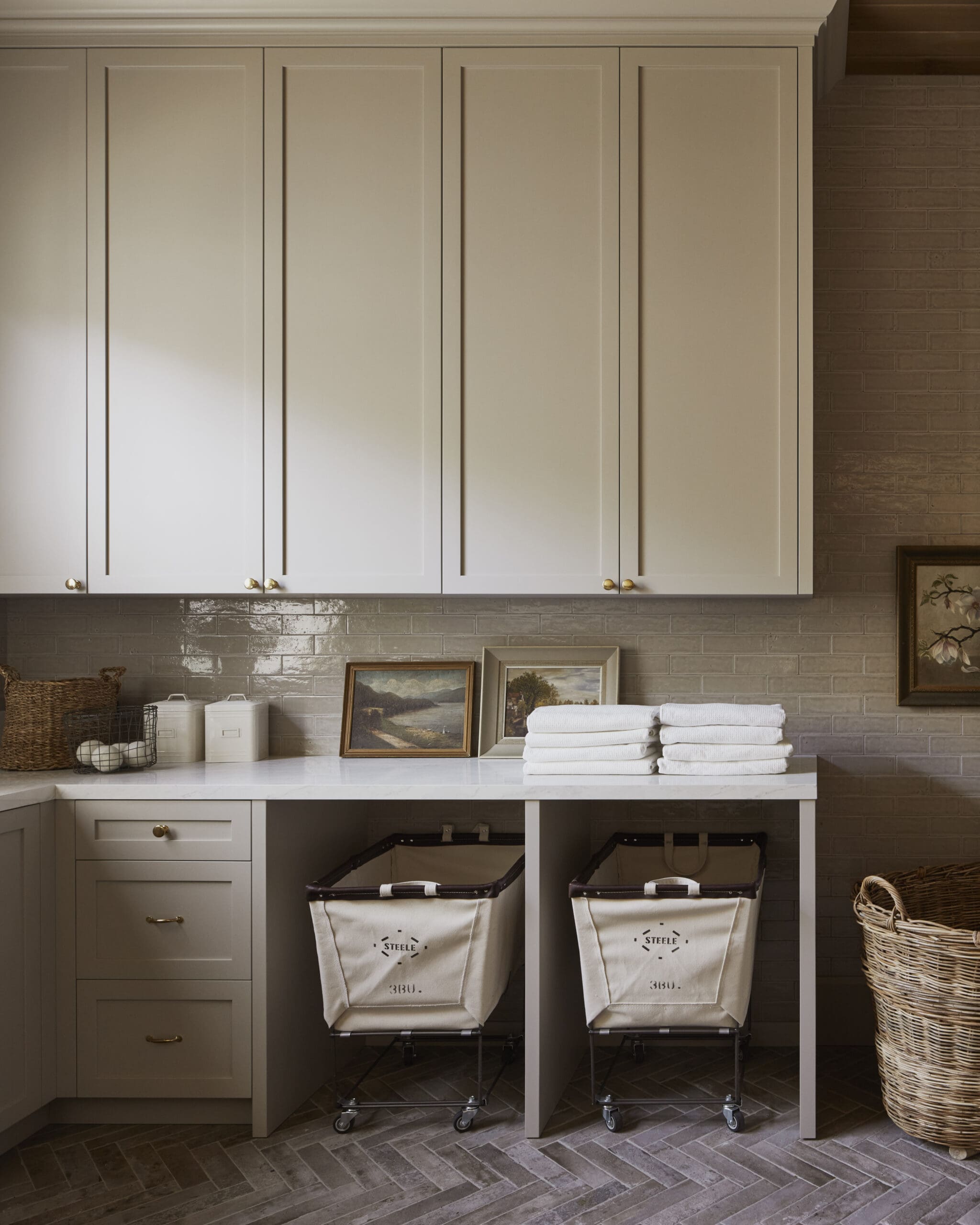 Beige kitchen cabinets above a white marble countertop with brick backsplash and two laundry hampers under a blue-gray table.