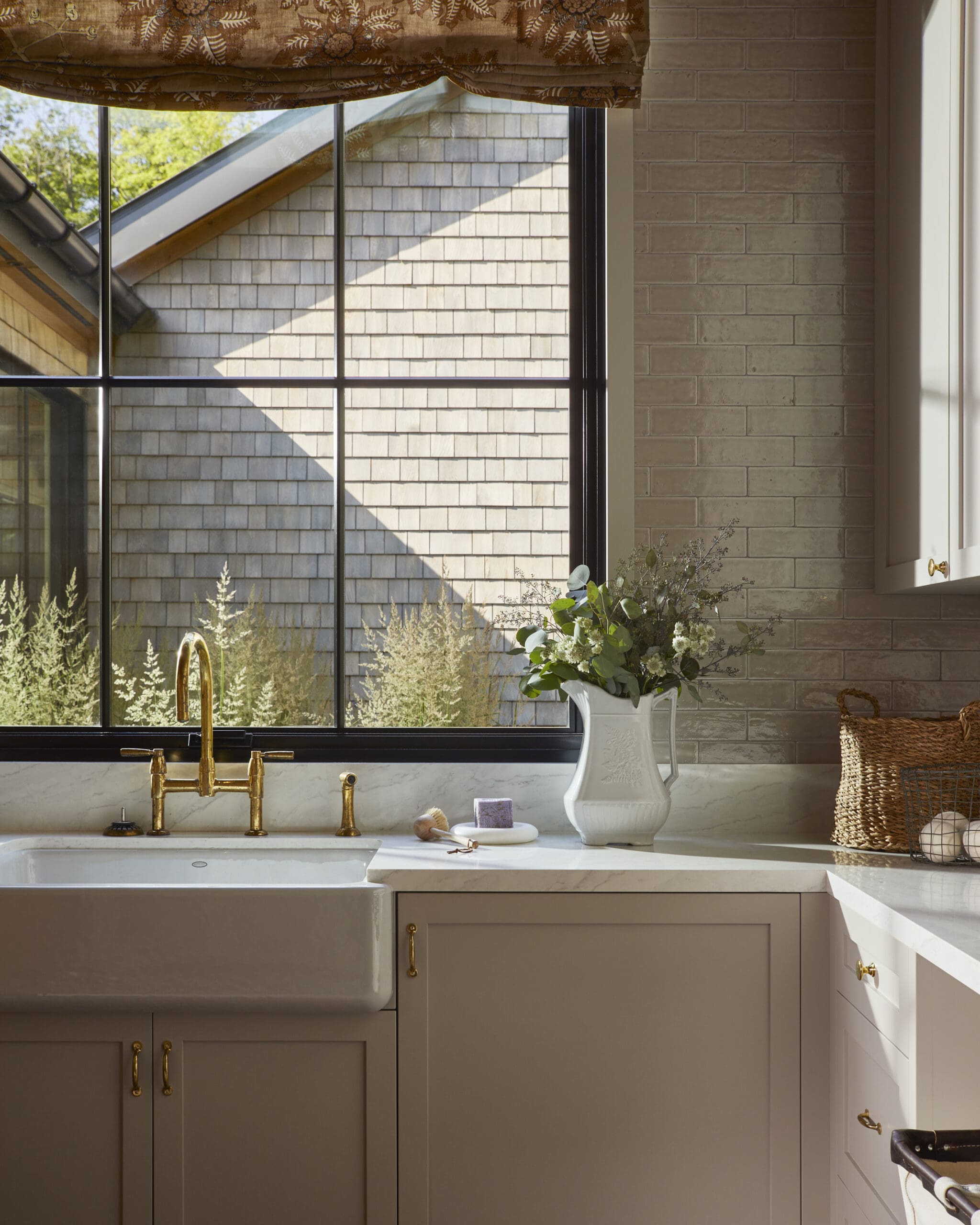 Kitchen sink area with a gold-toned faucet, white marble countertop, and a decorative vase of greenery near a large window showing an exterior shingled wall.