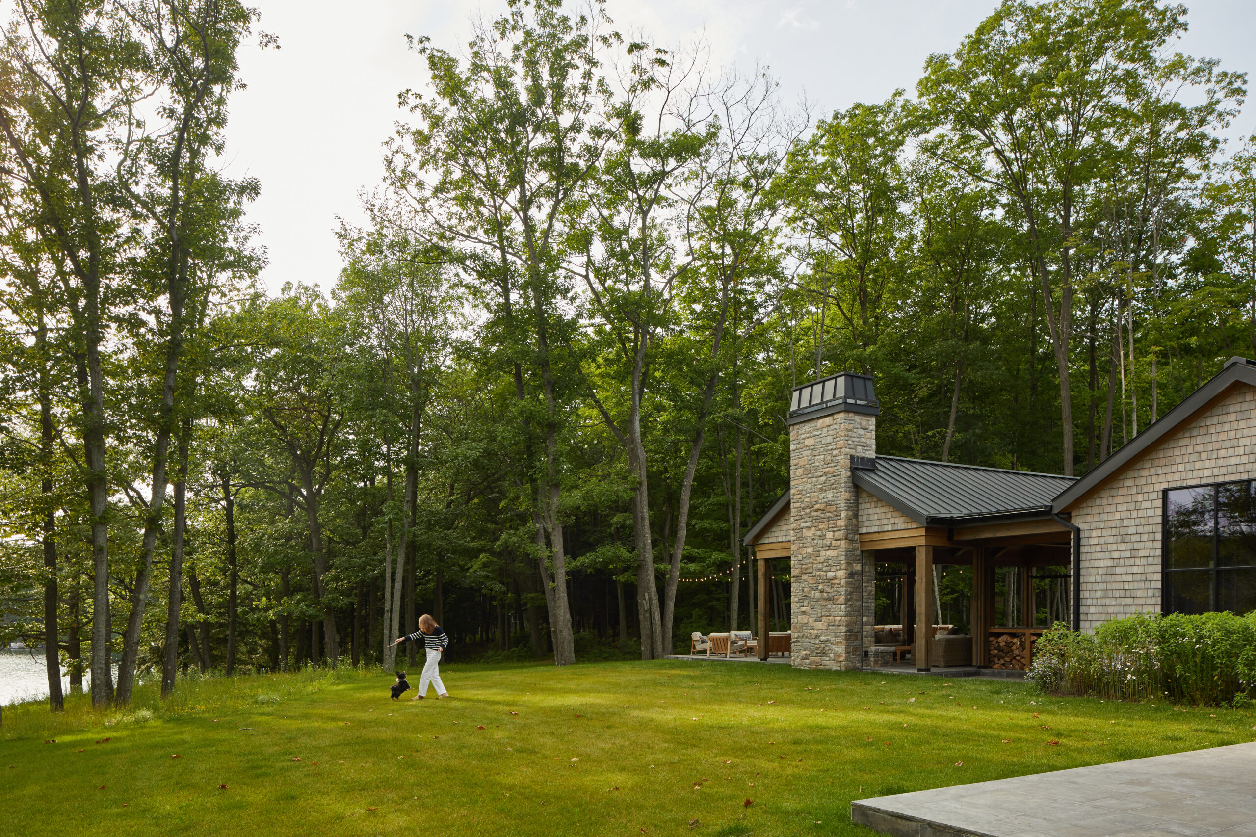 Woman walking a small dog across a green lawn in front of a brick house with a stone chimney and dense trees behind.