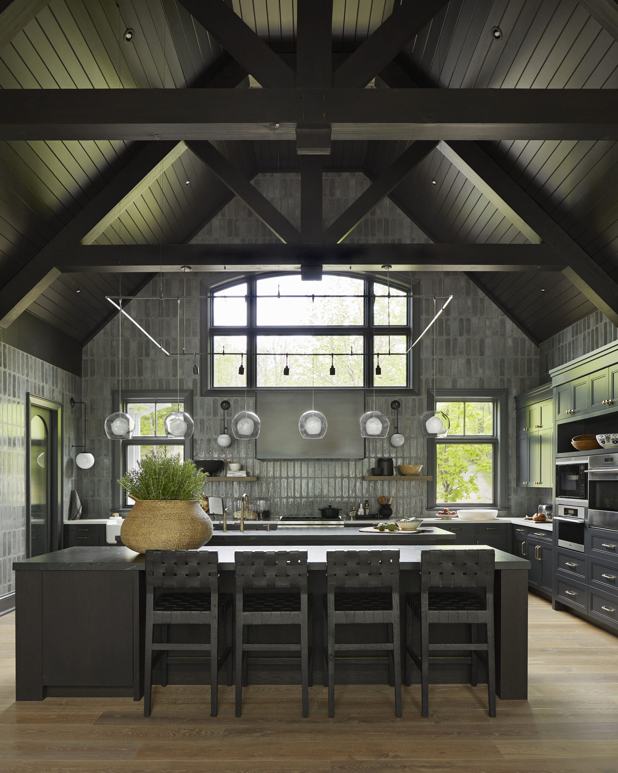 Dark kitchen with exposed wooden beams, grey tiled walls, and a large window above the sink area. A central black island with stools and a wicker planter on top creates a rustic-modern look.