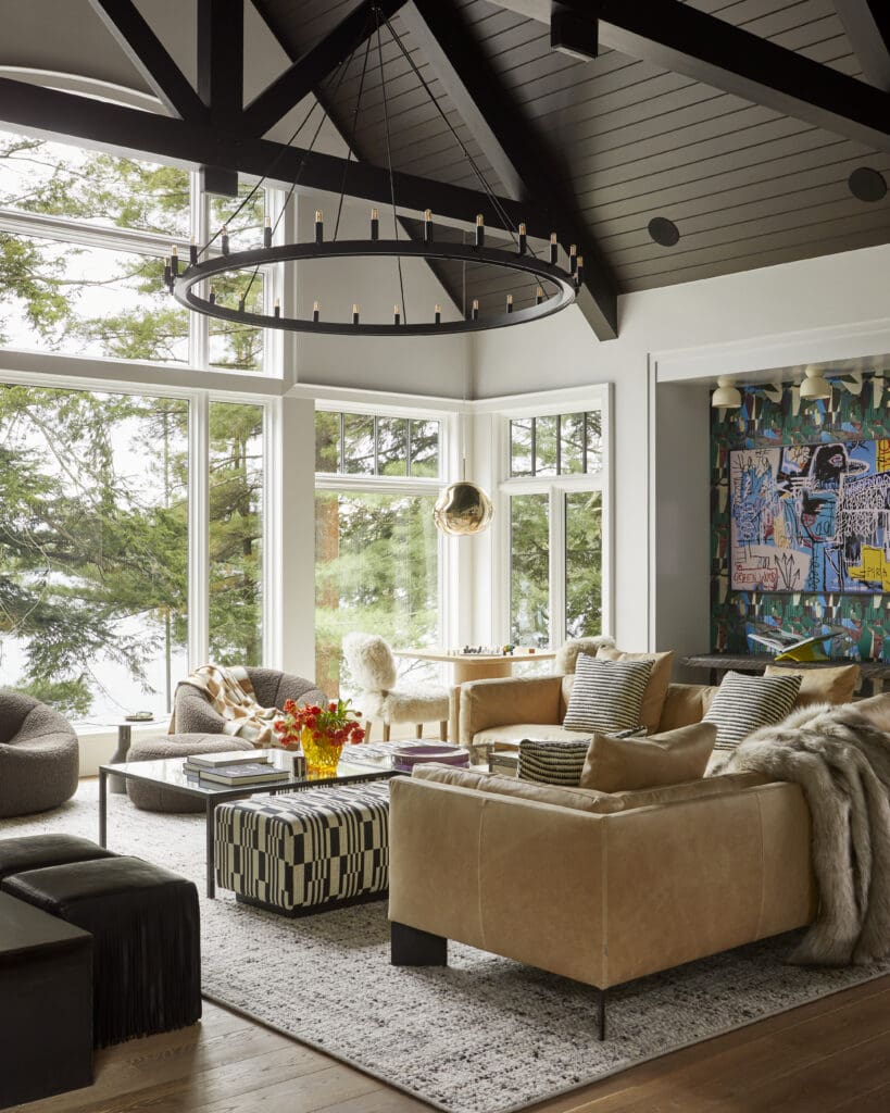 Open, sunlit living room with a large circular black chandelier, beige sofa, and a checkered ottoman by a glass coffee table.