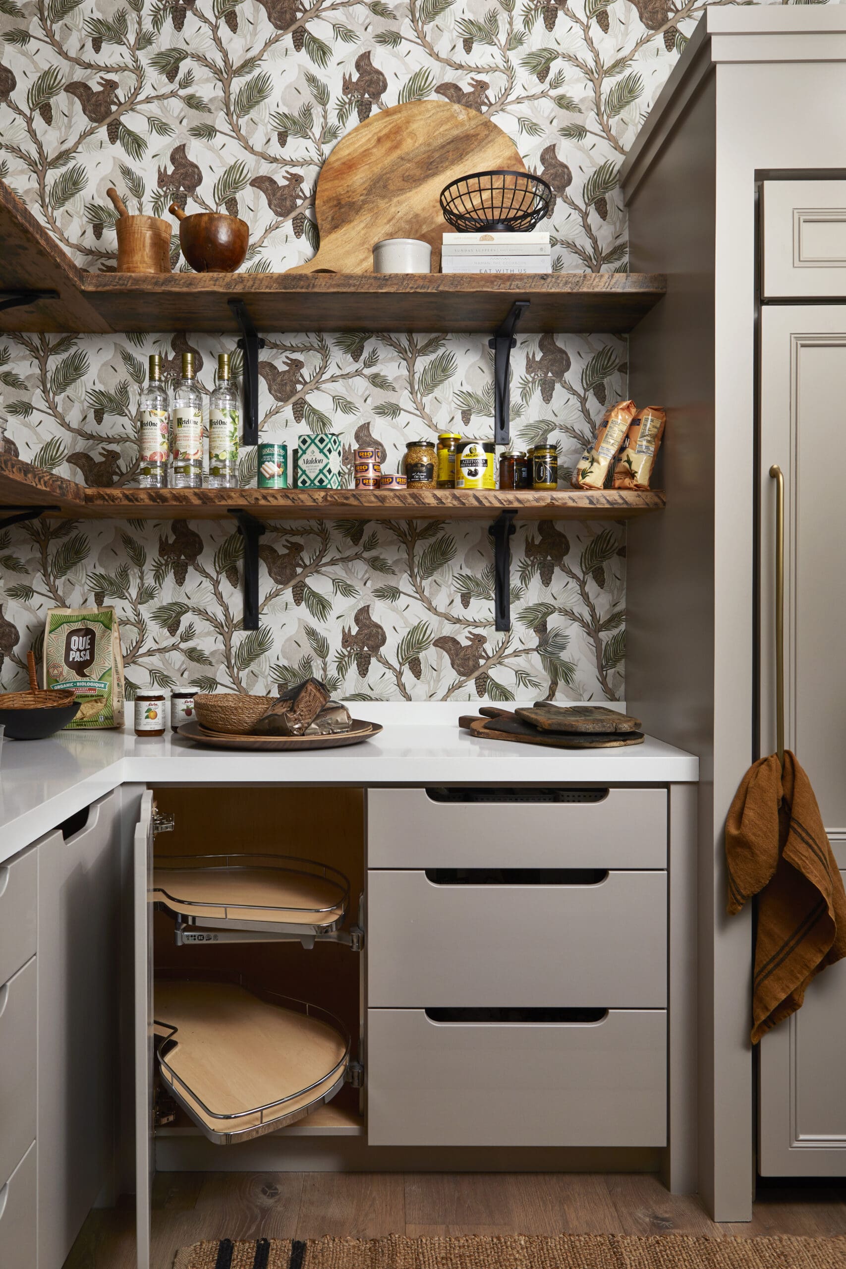 Open pantry with two wooden shelves against squirrel-pattern wallpaper; bottles, jars, cans, and bread arranged on the shelves.