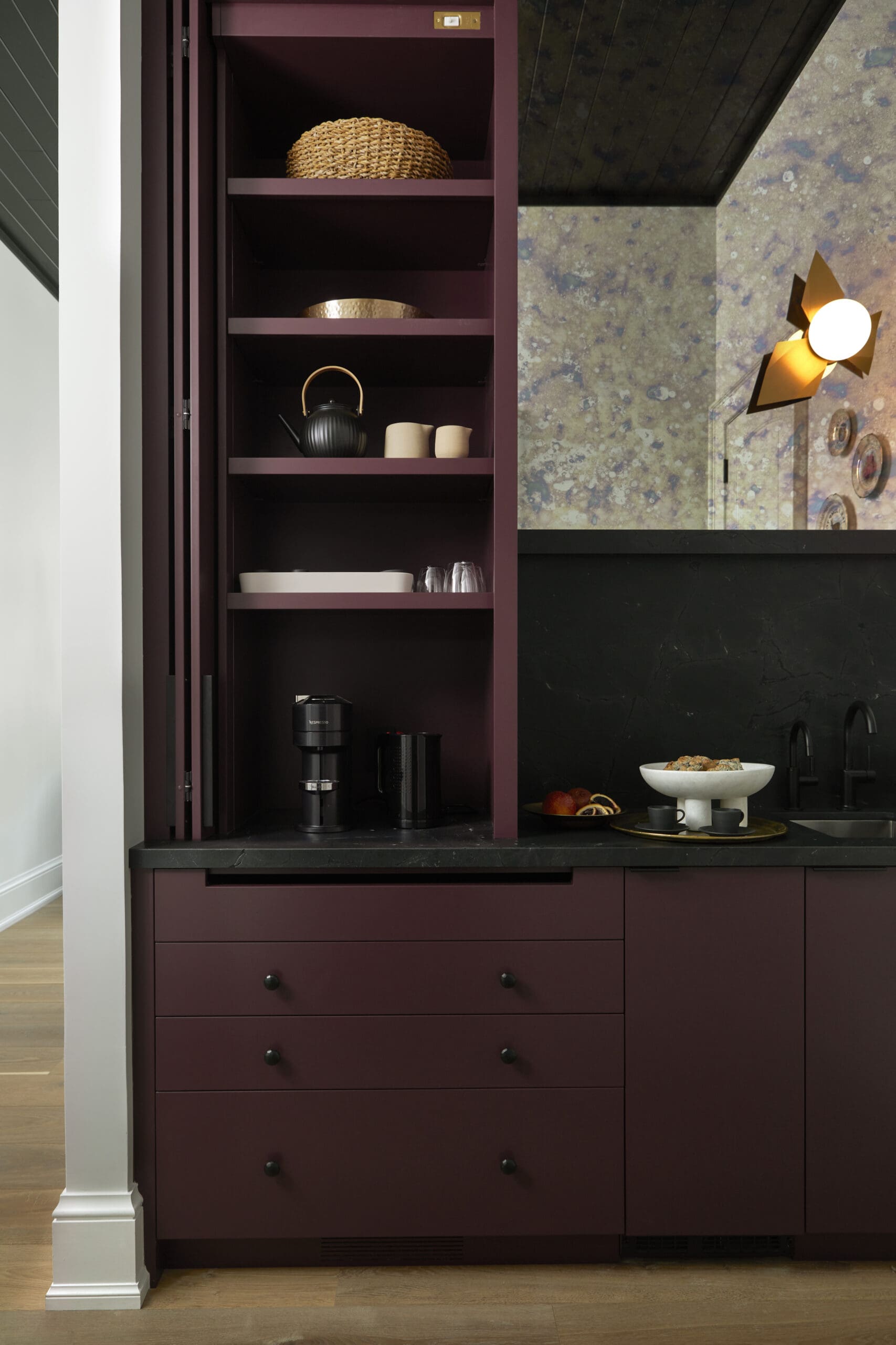 Maroon kitchen cabinet with open shelves displaying a teapot, cups, and a woven basket; pastry stand on the counter nearby.