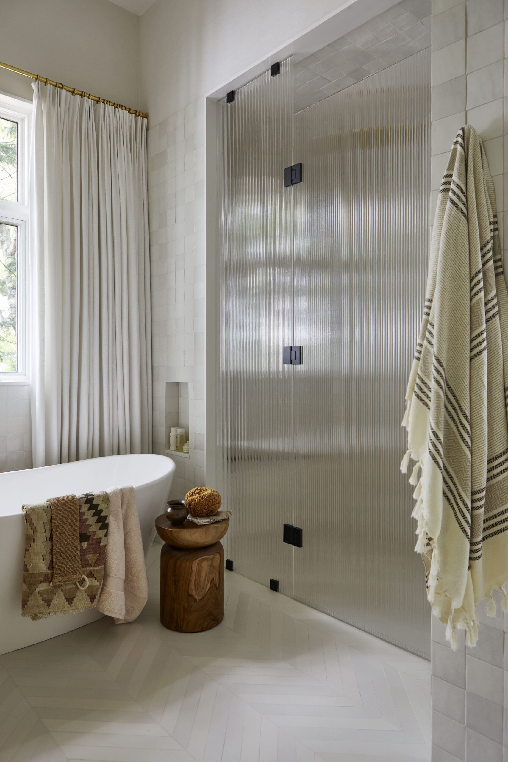 White freestanding bathtub by a window with white curtains, a wooden stool holding towels and bath items, and a ribbed glass shower enclosure in a neutral bathroom.