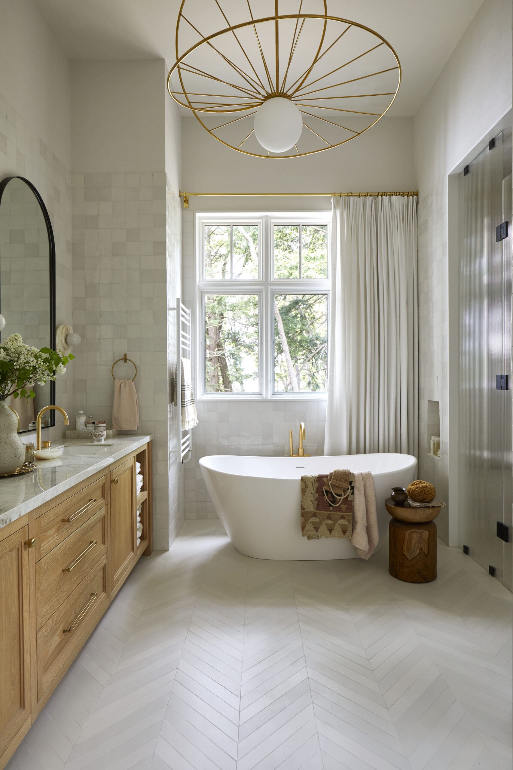 Bright bathroom with a freestanding white tub, gold fixtures, and a large window framed by white curtains.