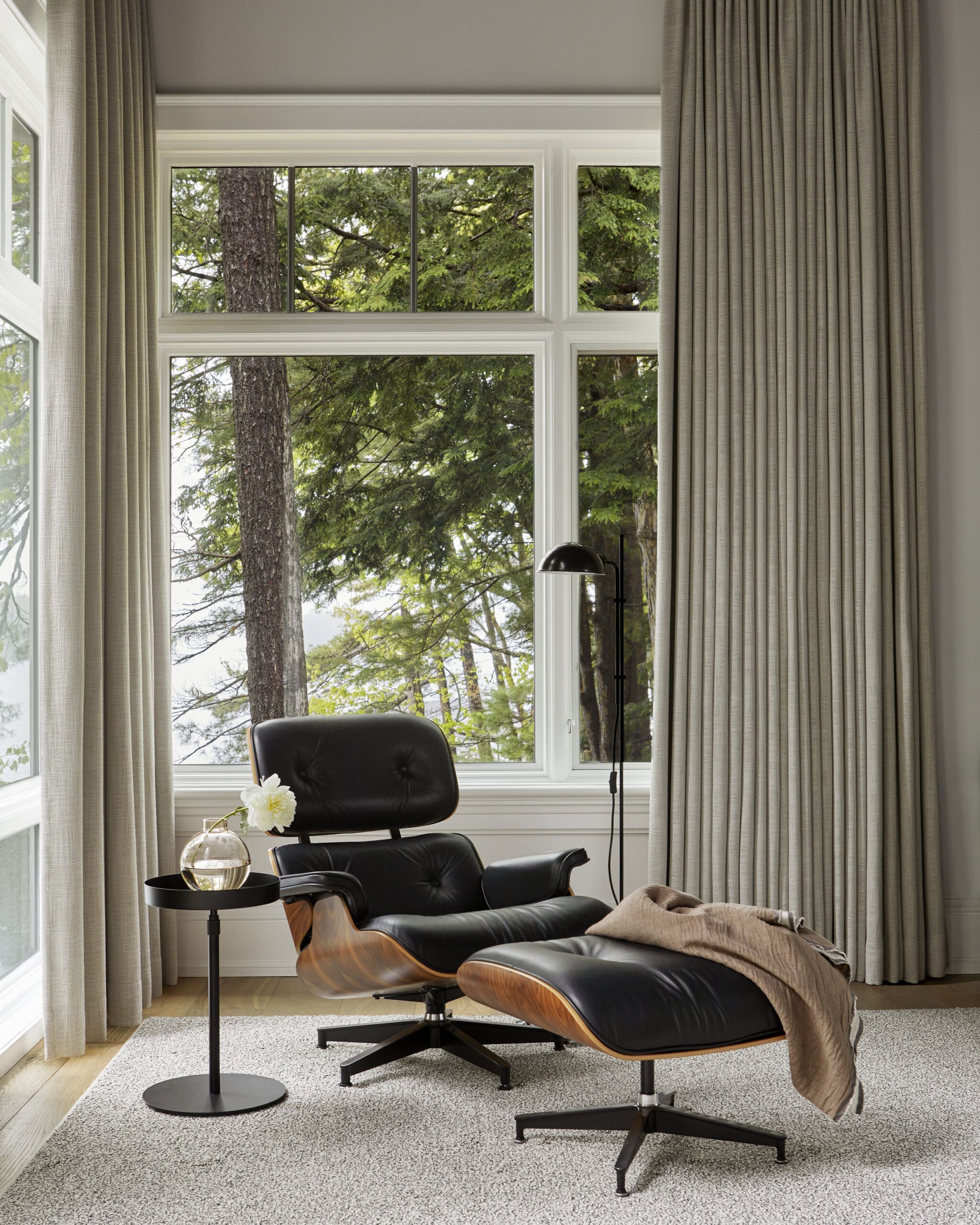 Black leather Eames lounge chair with matching ottoman, wooden base, beside a round side table with a glass vase and white flower in a bright living corner by a large window with gray drapes.