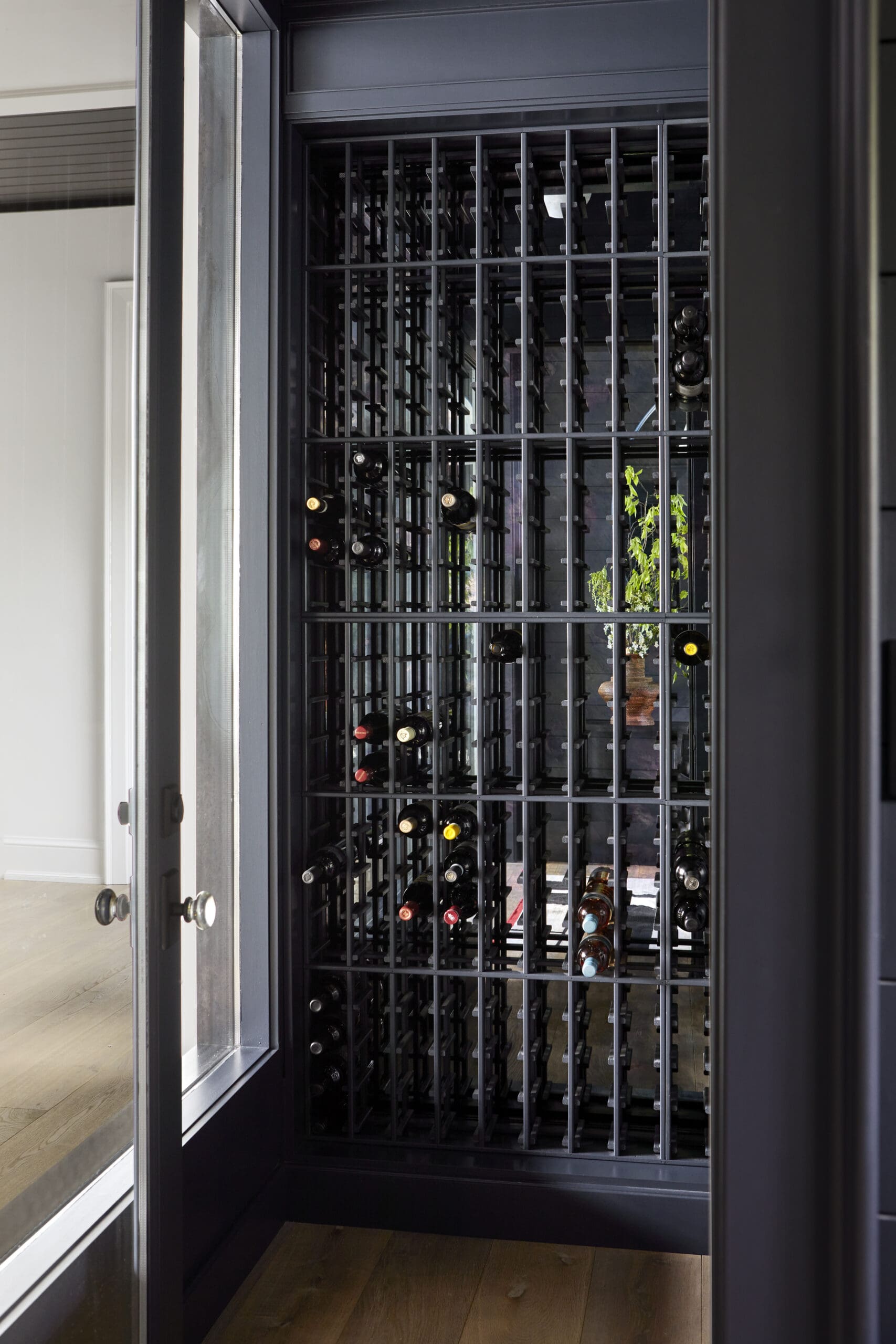 Open glass-front wine cooler with grid shelves filled with wine bottles inside a modern room.
