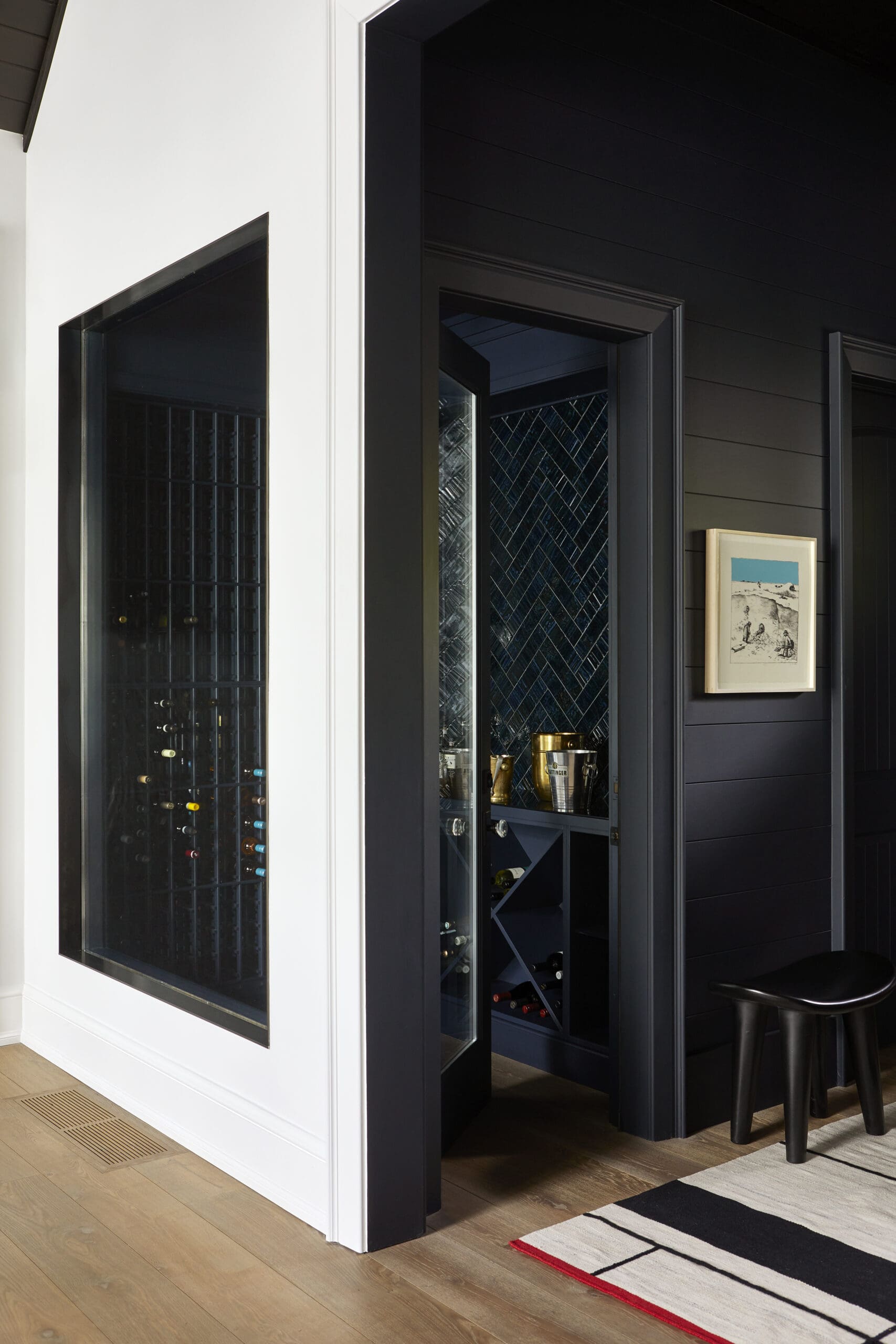 White-framed glass-front wine cabinet built into a white wall, showing bottles inside, with a dark wall cabinet and a small framed picture on the wall.