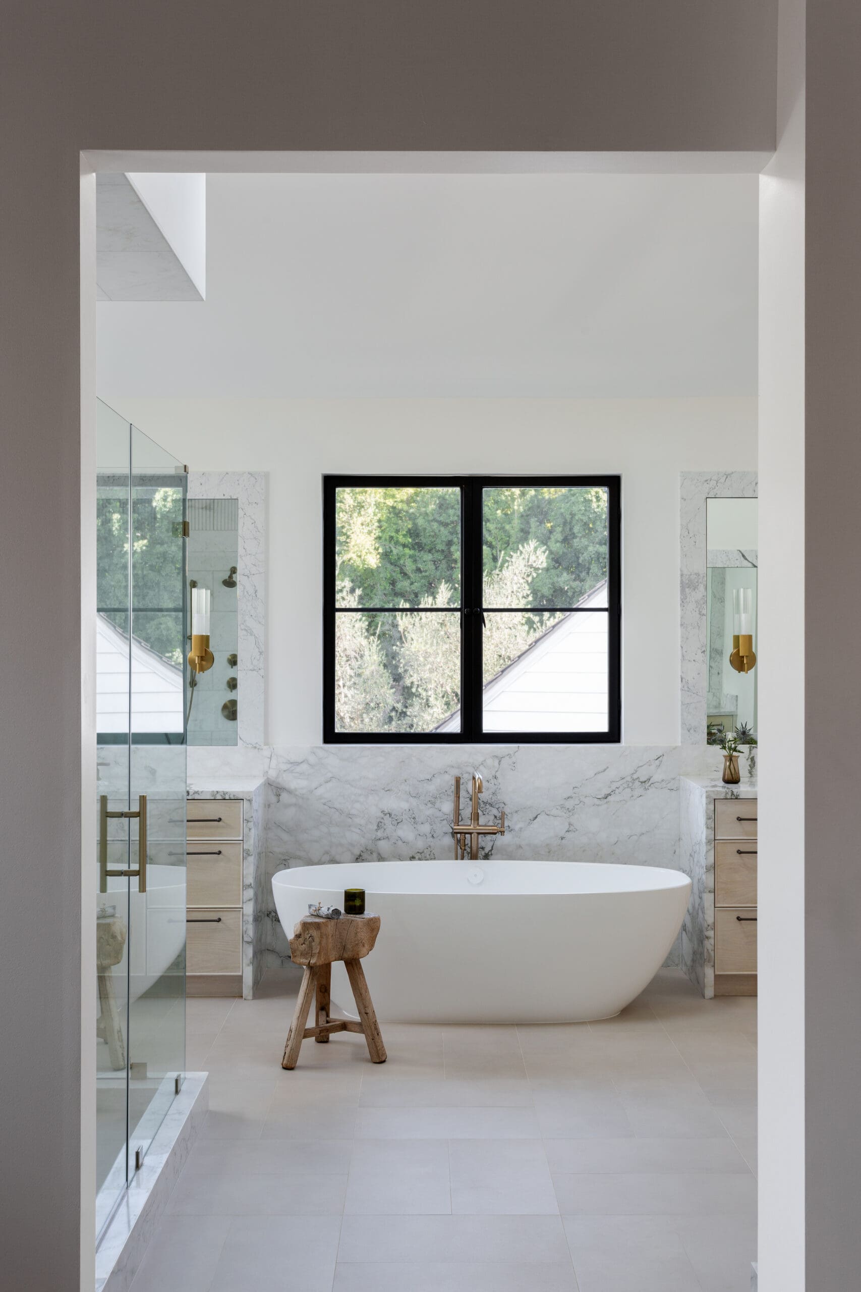 Modern bathroom with a white freestanding bathtub, marble surfaces, and a large black-framed window.