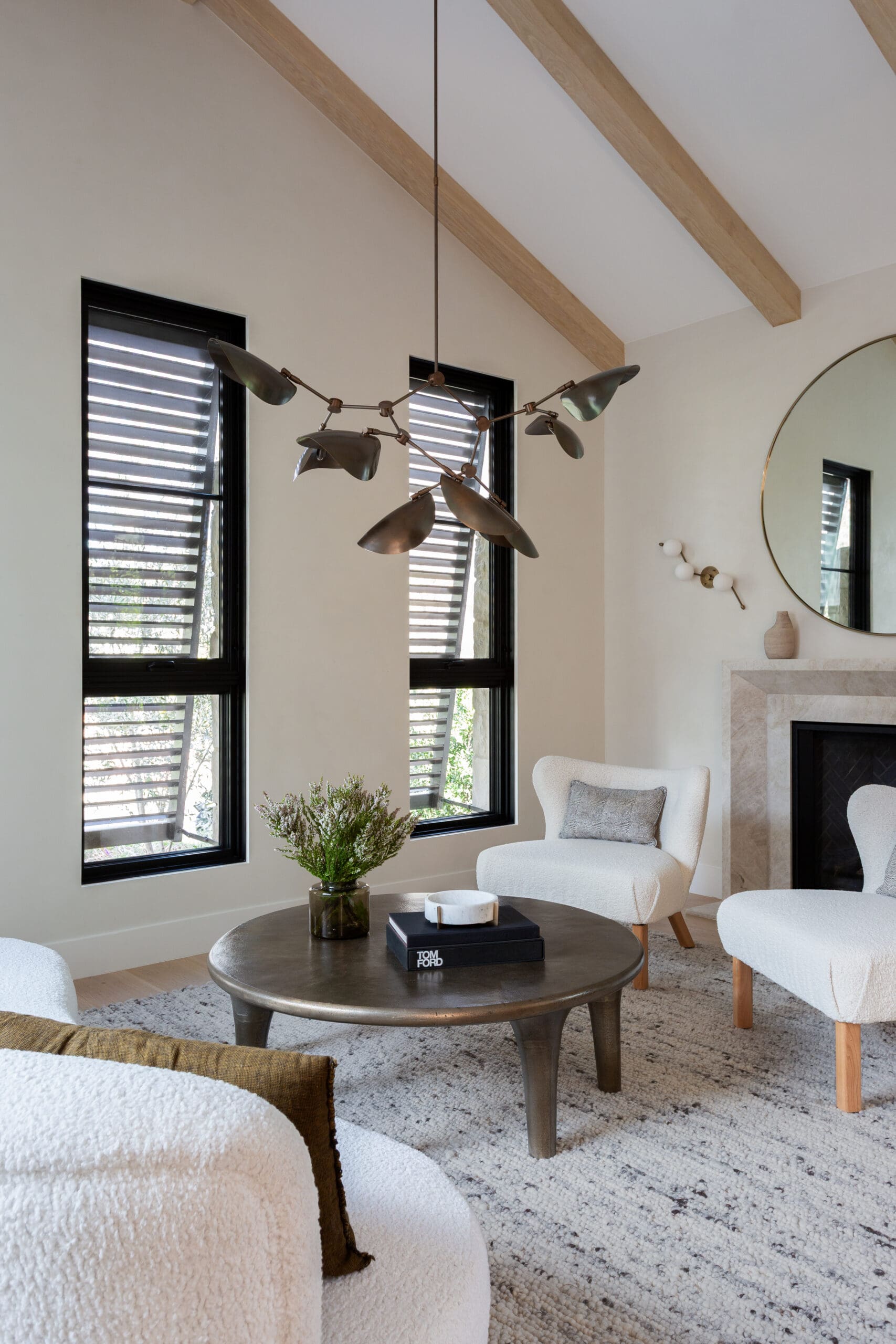 Bright living room with white boucle chairs, a round coffee table, and a modern sculptural chandelier; large black-framed windows with shutters behind.