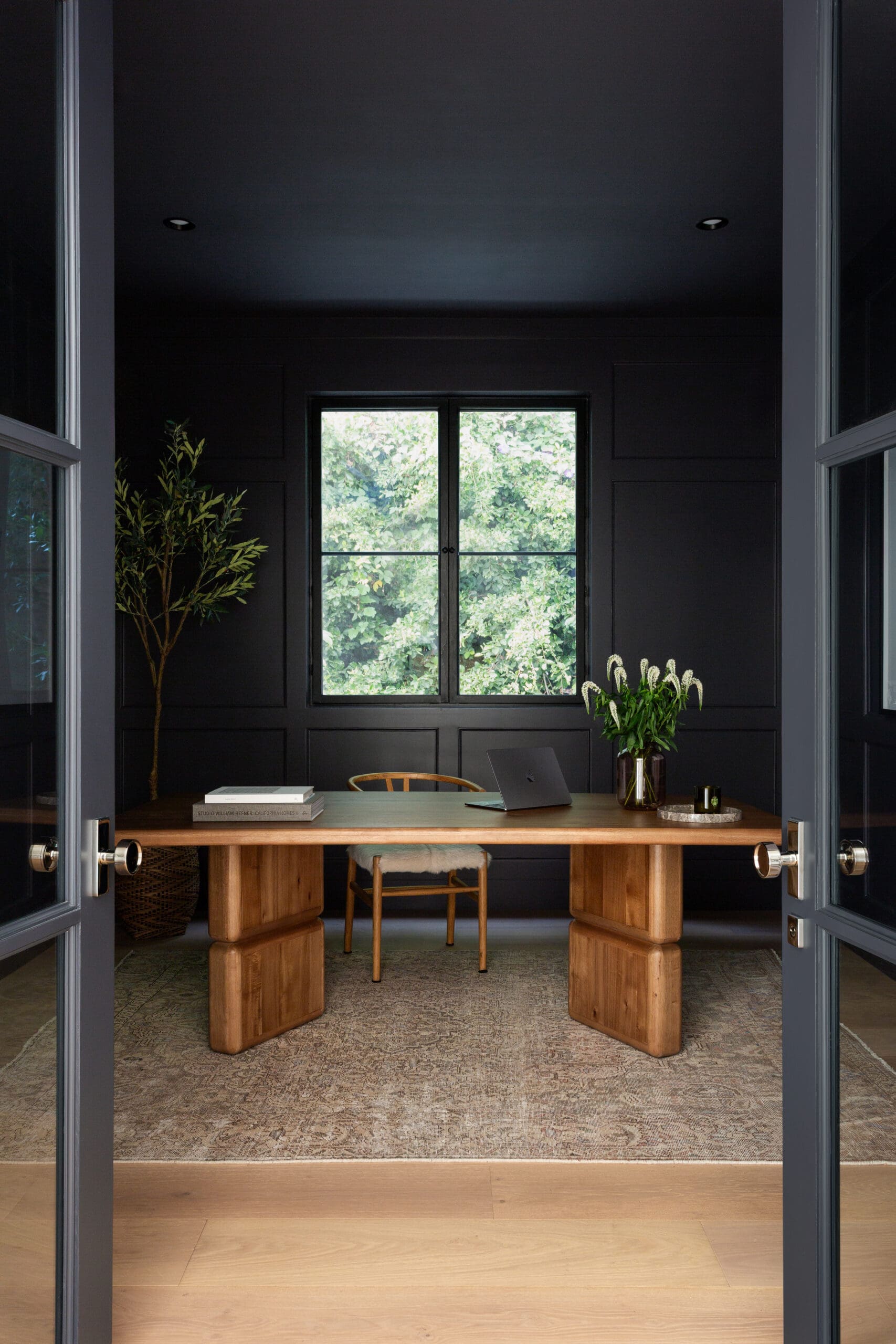 Open double doors reveal a dark-paneled office with a wooden desk, laptop, and green plants, plus a window showing trees outside.