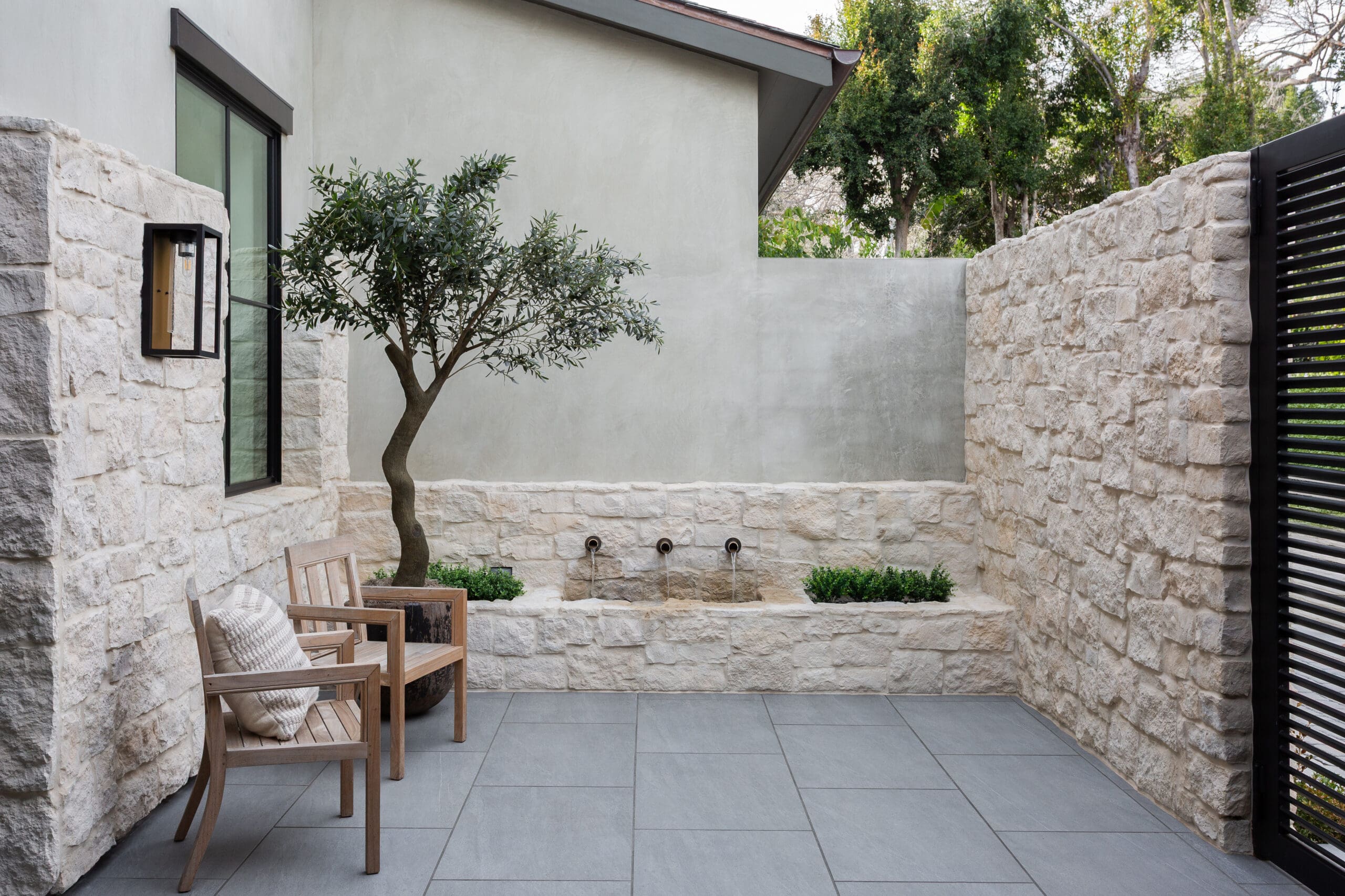 Stone-walled courtyard with a small olive tree, wooden chairs, and cushioned seating near a lantern-style wall light.