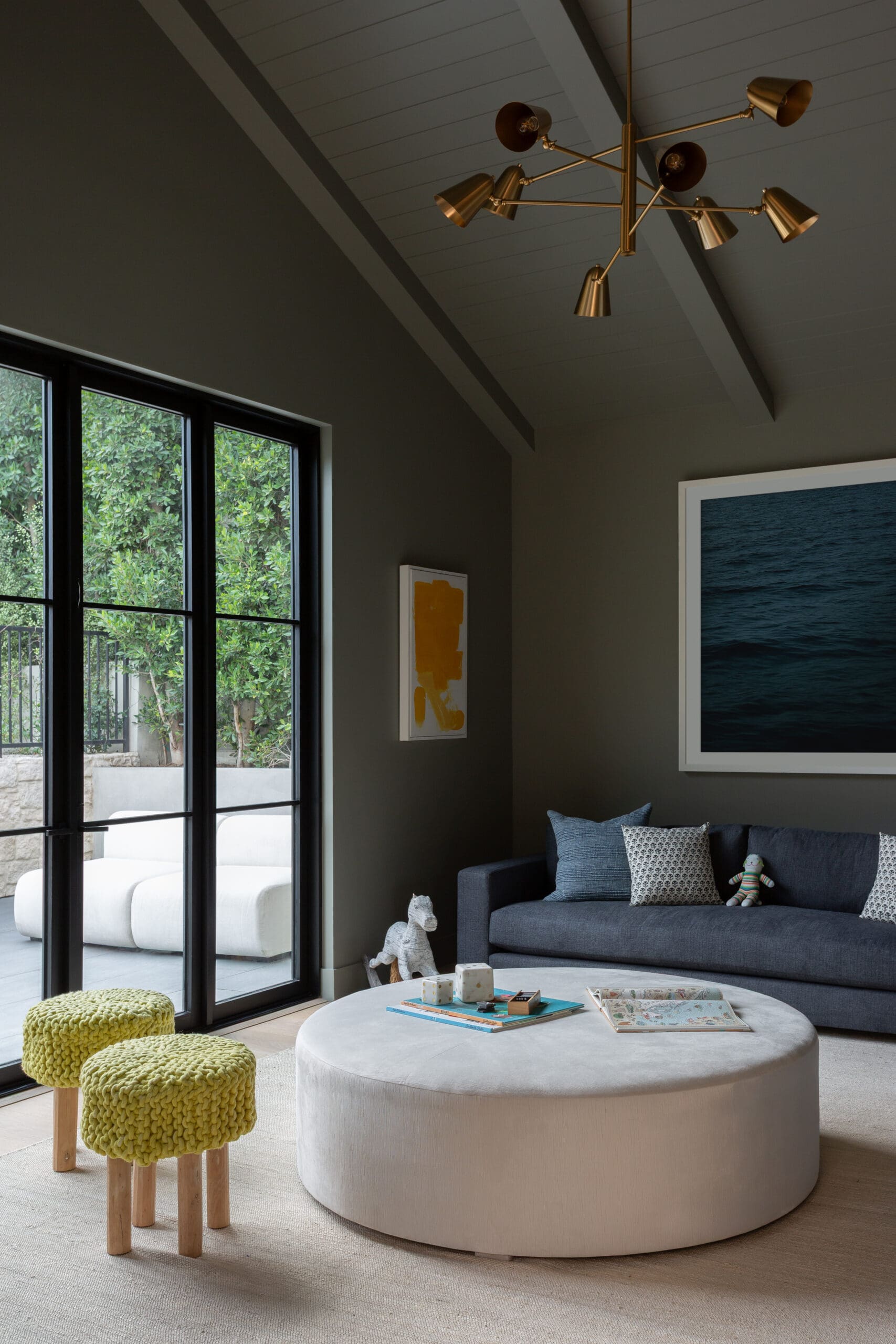 Modern living room with dark gray walls, black-framed glass doors, a gray sofa, and a large round beige ottoman; two bright yellow knitted stools nearby.
