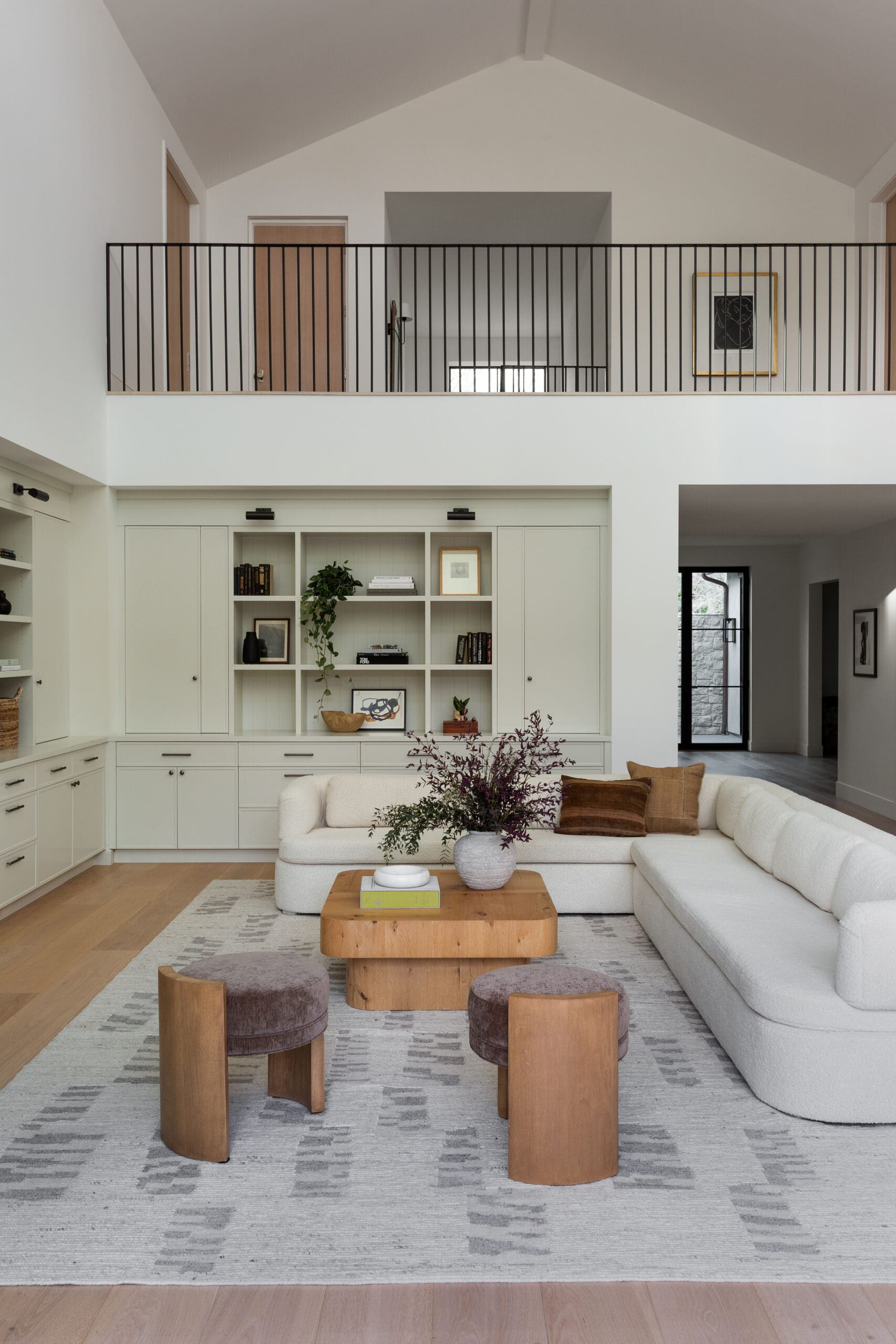 Bright two-story living room with white built-in cabinets, a white sectional sofa, and a natural wood coffee table on a light gray patterned rug.