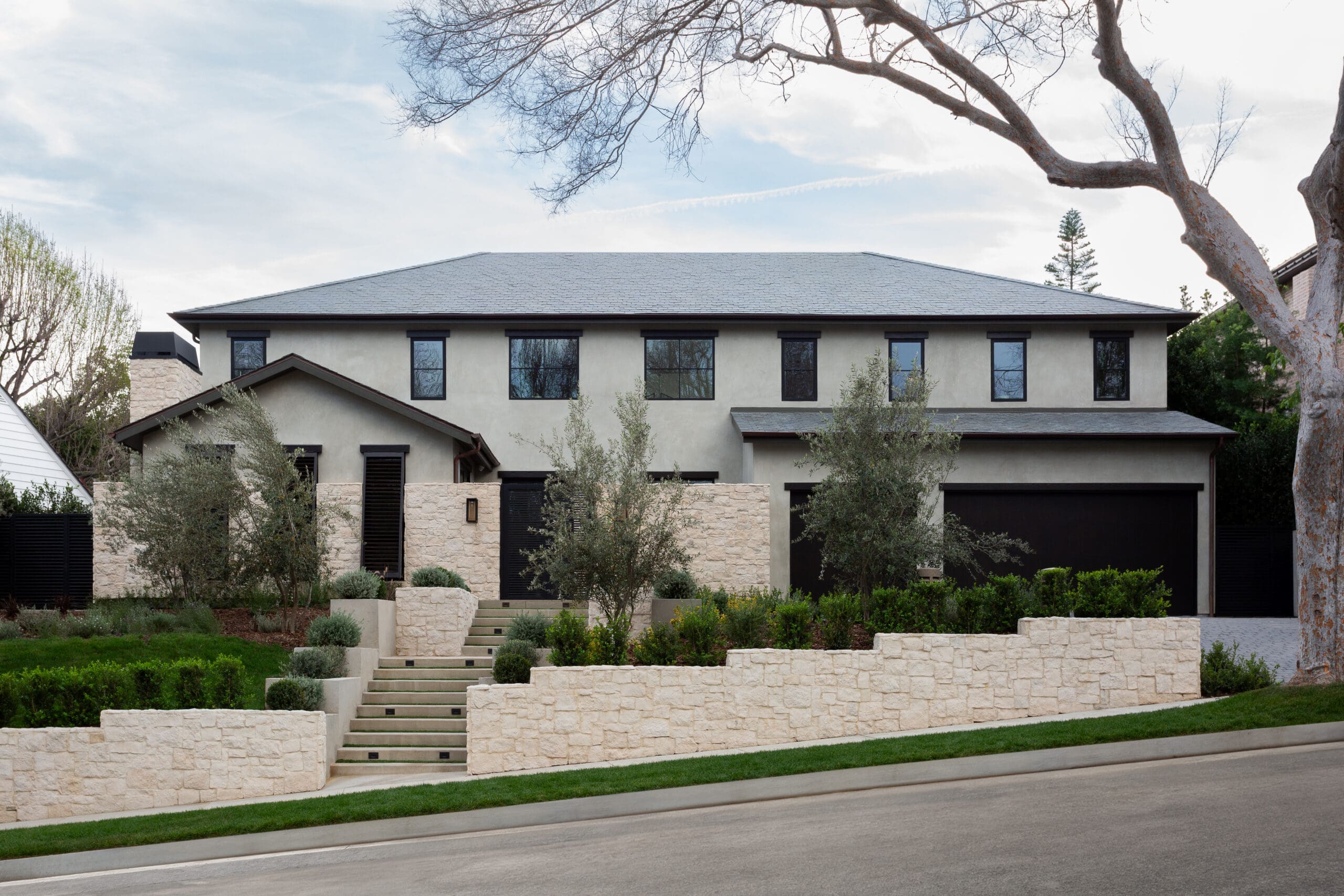Modern beige two-story house with black-framed windows, stone retaining walls, and manicured landscaping as a front yard.