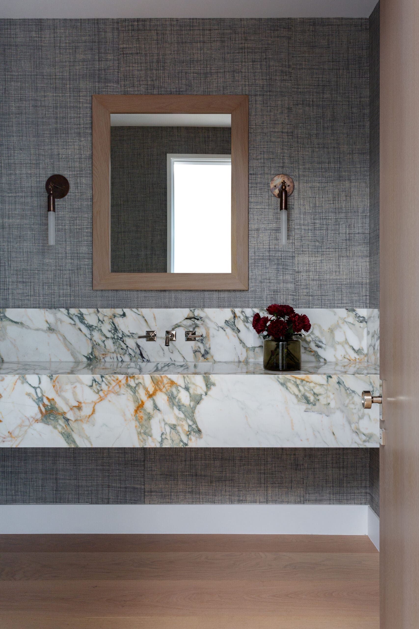 Wood-framed rectangular mirror above a marble bathroom countertop with two copper wall sconces and a vase of red flowers on a blue textured wall.