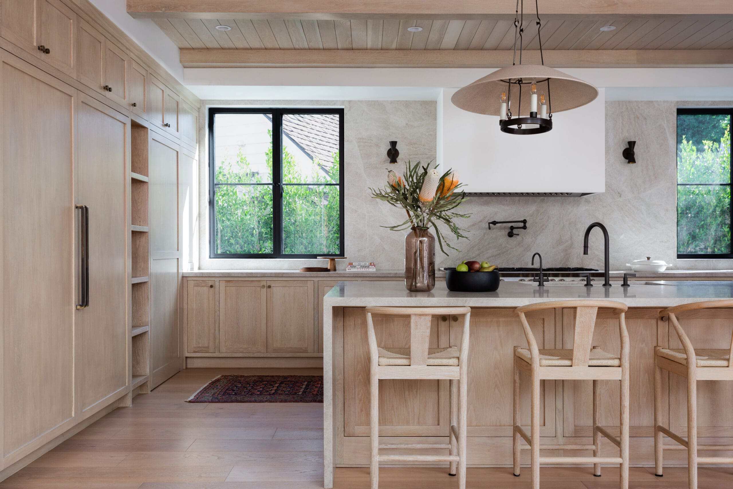Bright, oak-toned kitchen with a large island, bar stools, and black-framed windows showing greenery outside.