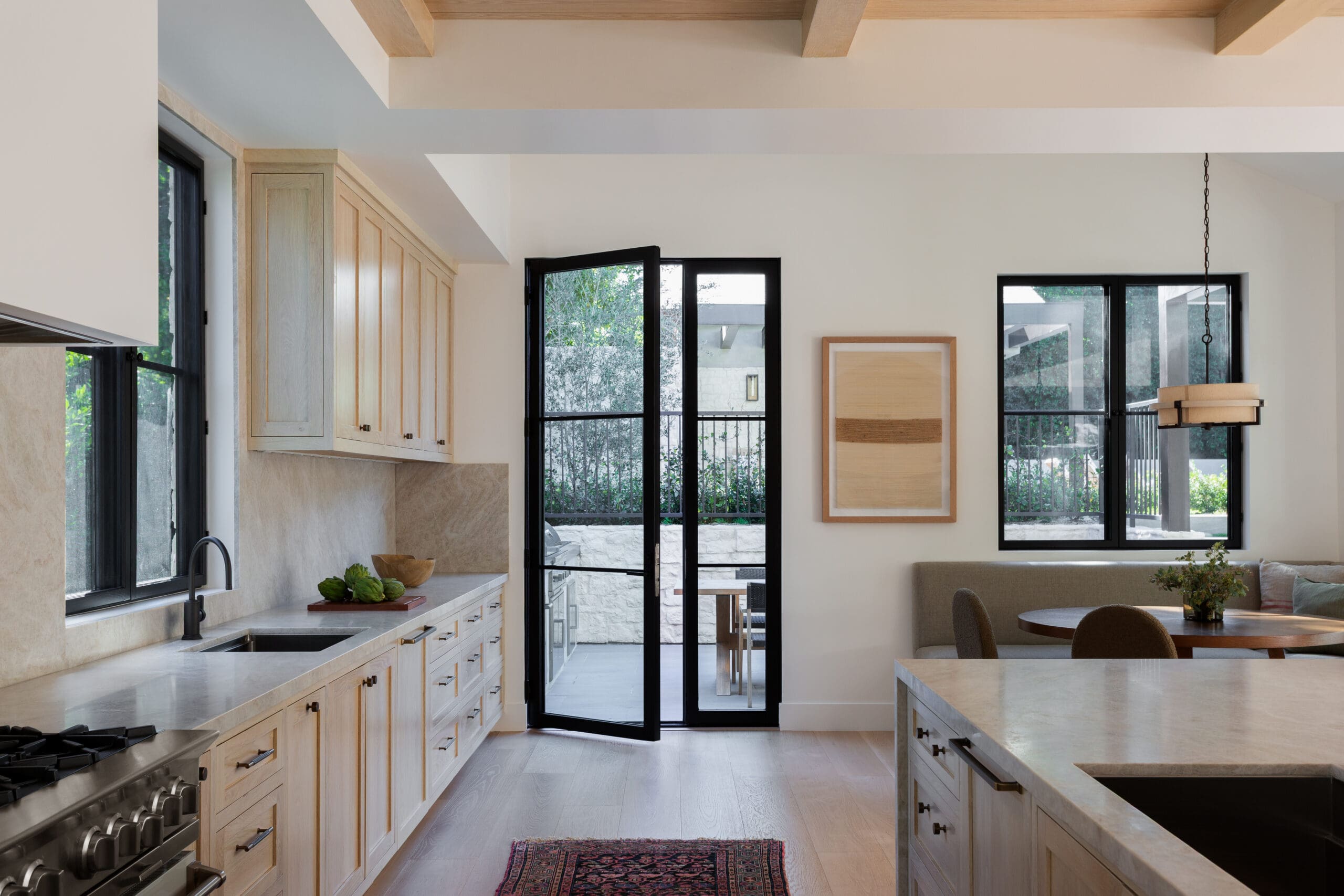 Bright open-concept kitchen with light wood cabinets, marble countertops, and a black-framed glass door leading to an outdoor patio.