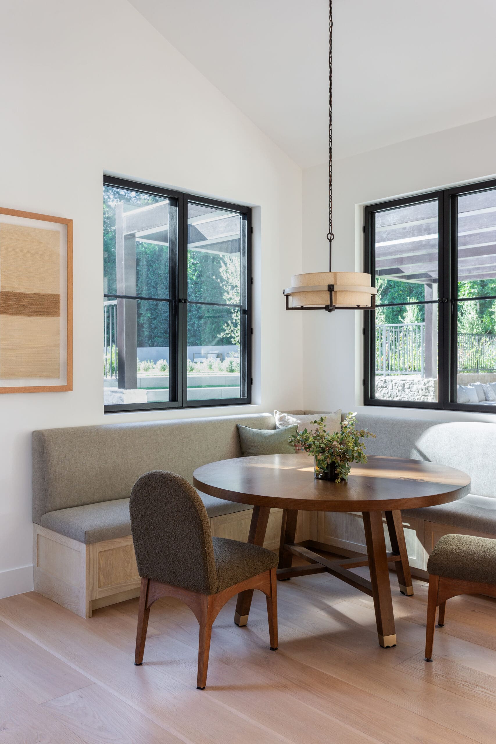 Cozy dining nook with a round wooden table, built‑in beige bench, two chairs, and a hanging pendant light by large black-framed windows.