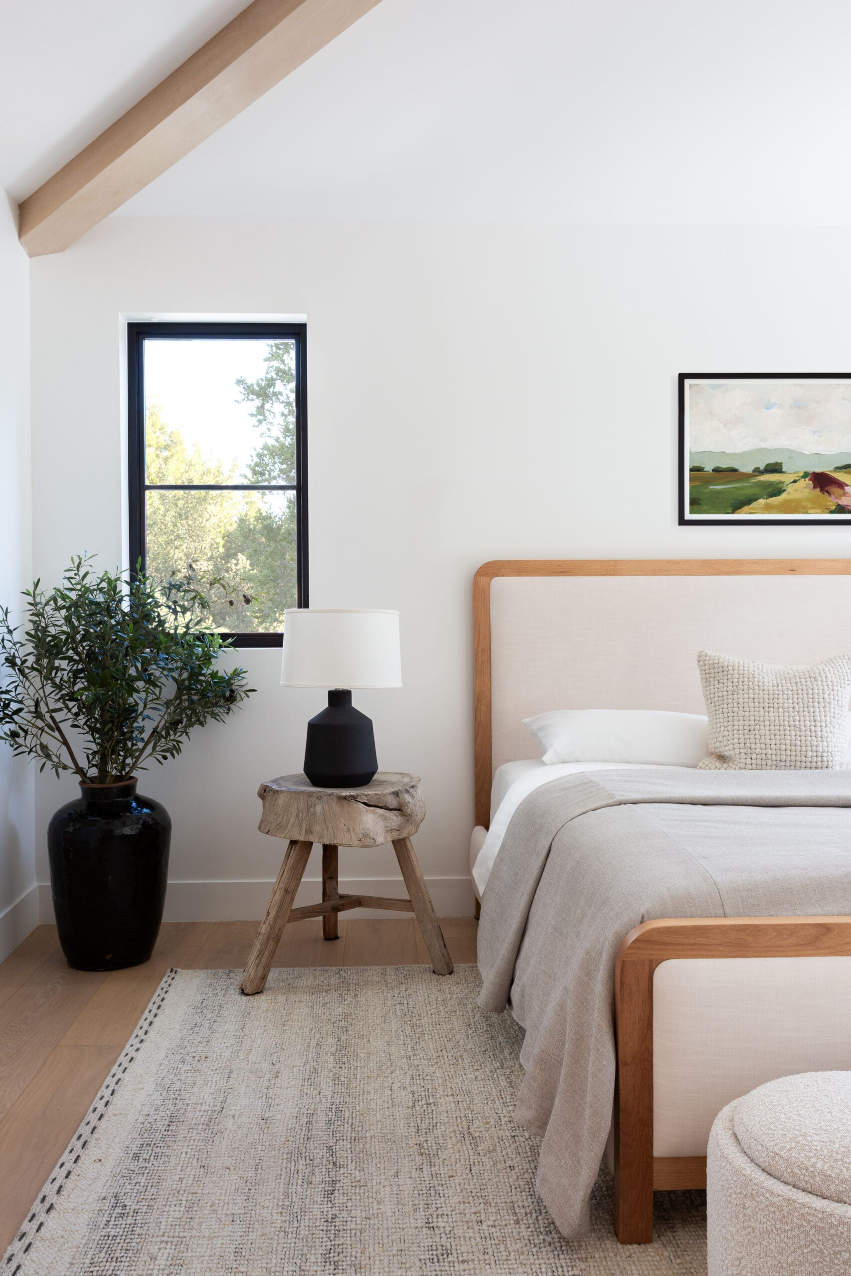 Bright bedroom with a light wood bed frame, beige linens, a black bedside lamp on a rustic stump side table, and a large window with greenery outside.",